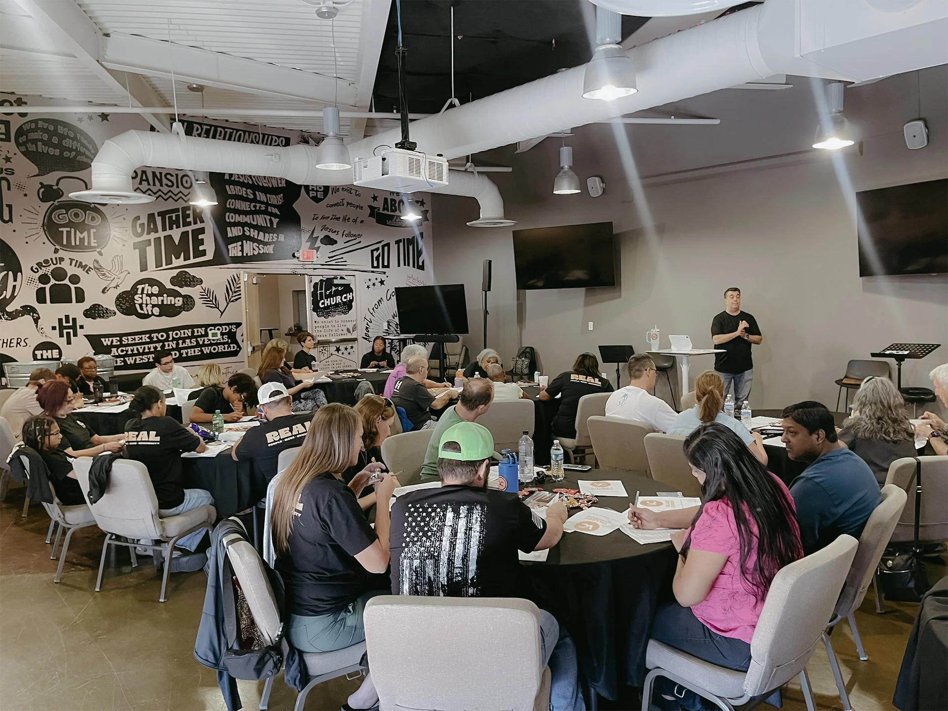 Indoor conference or seminar room with people seated at round tables listening to a speaker at the front. The room has a gray wall, several ceiling lights, and a wall mural with religious and inspirational messages.
