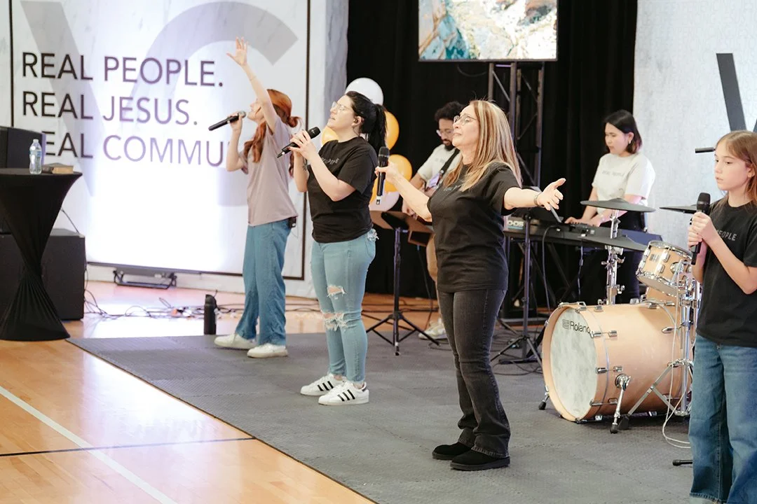 Group of people on stage leading worship with microphones, a drummer in the background, and a large screen displaying Christian messages.