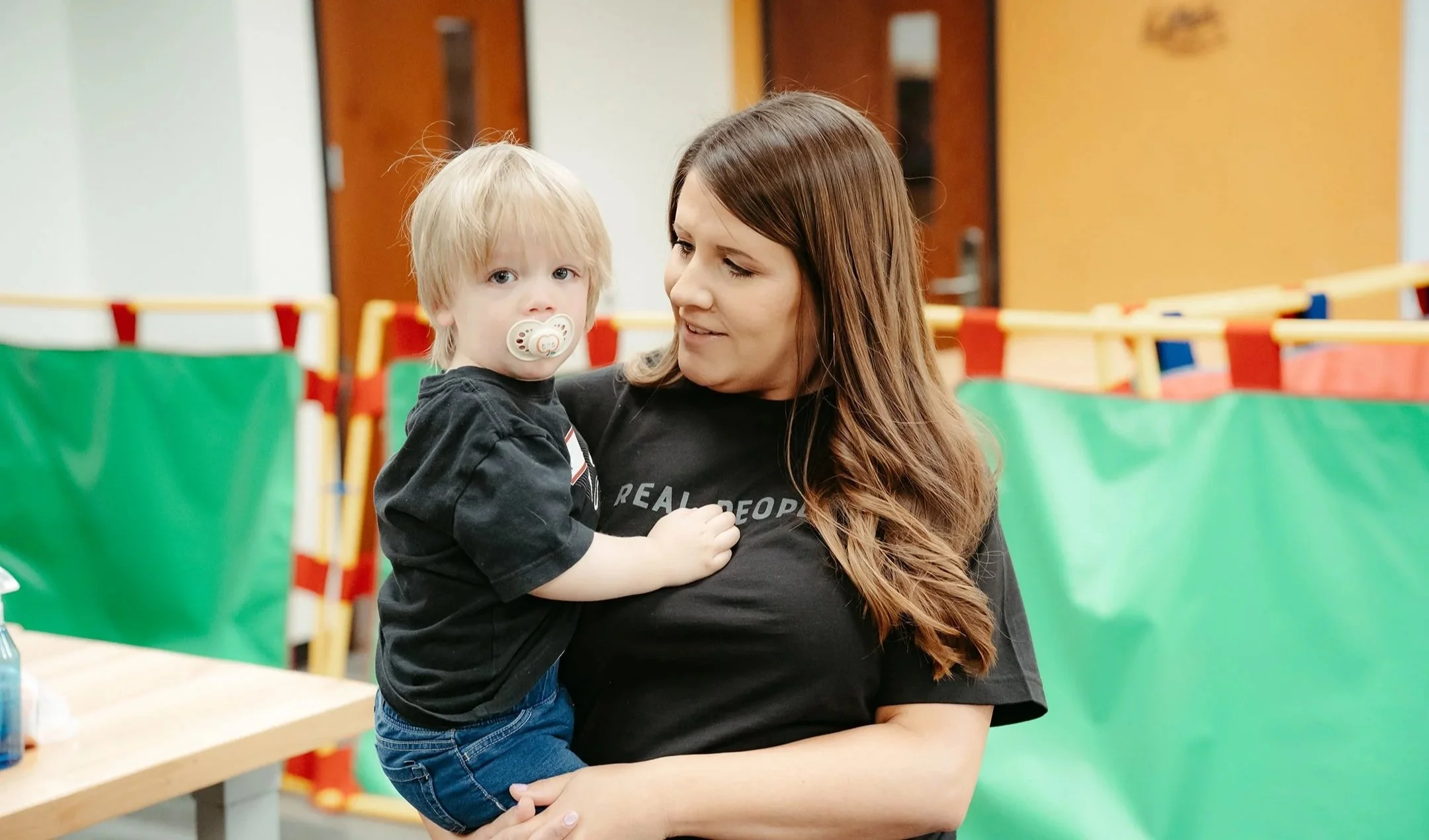 A woman with long brown hair holding a young boy with a pacifier in his mouth. The boy has blonde hair and is wearing a black t-shirt and jeans. The background shows a room with colorful barriers.