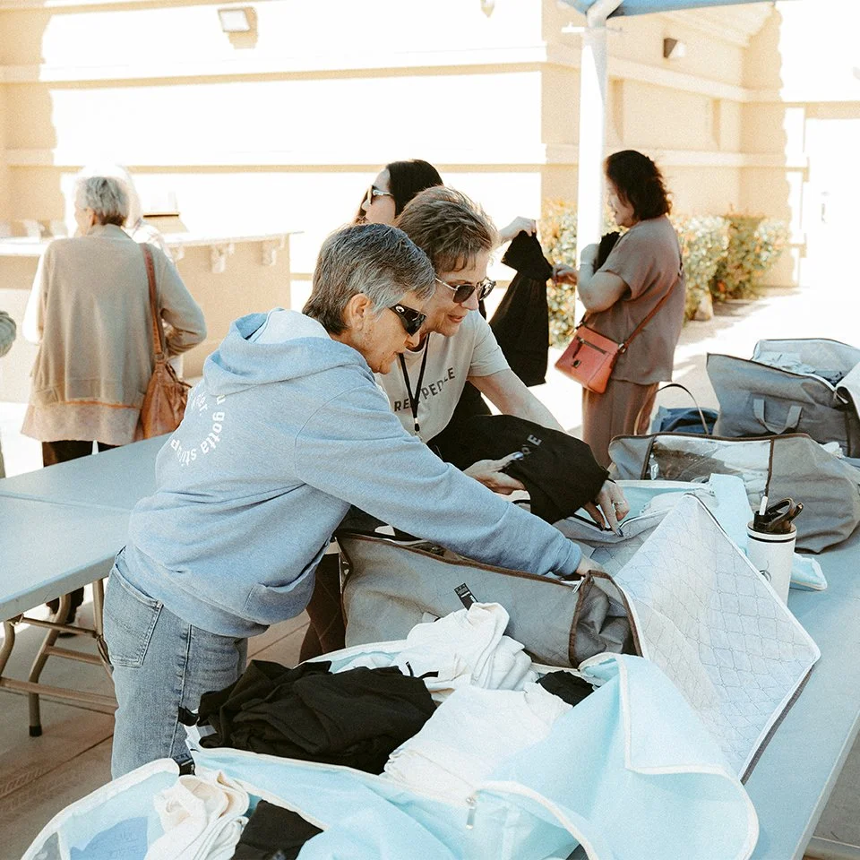Two women looking through luggage and clothes on a table at an outdoor location with other people in the background.