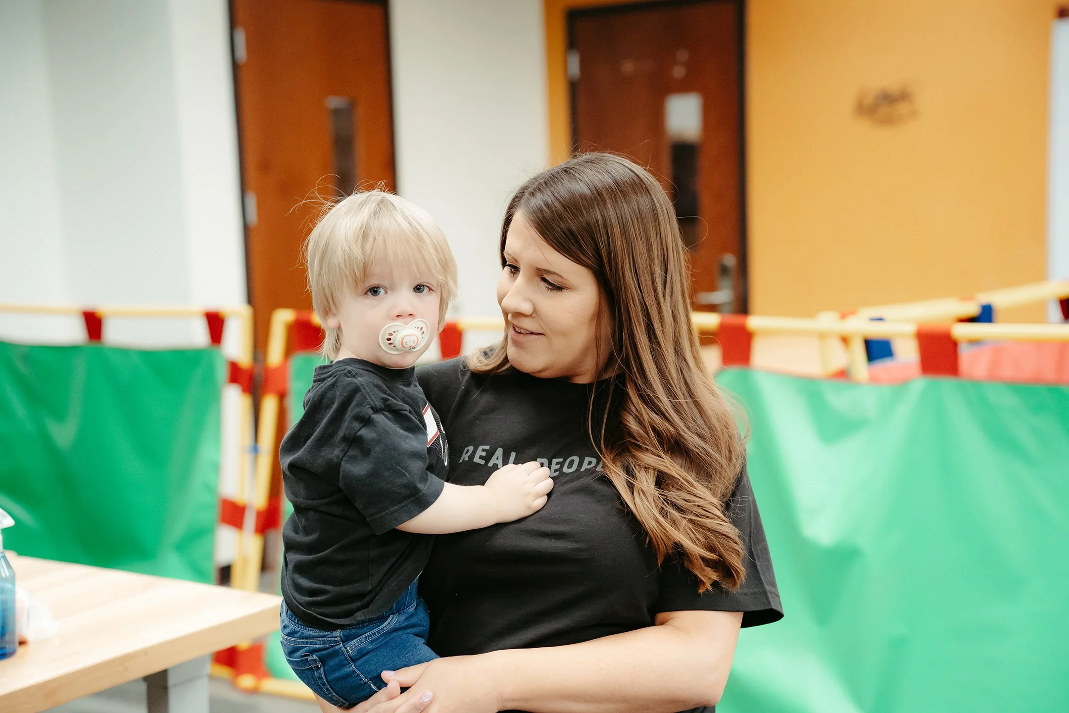 A woman holding a young boy with a pacifier in his mouth in a room with colorful barriers and wooden doors in the background.