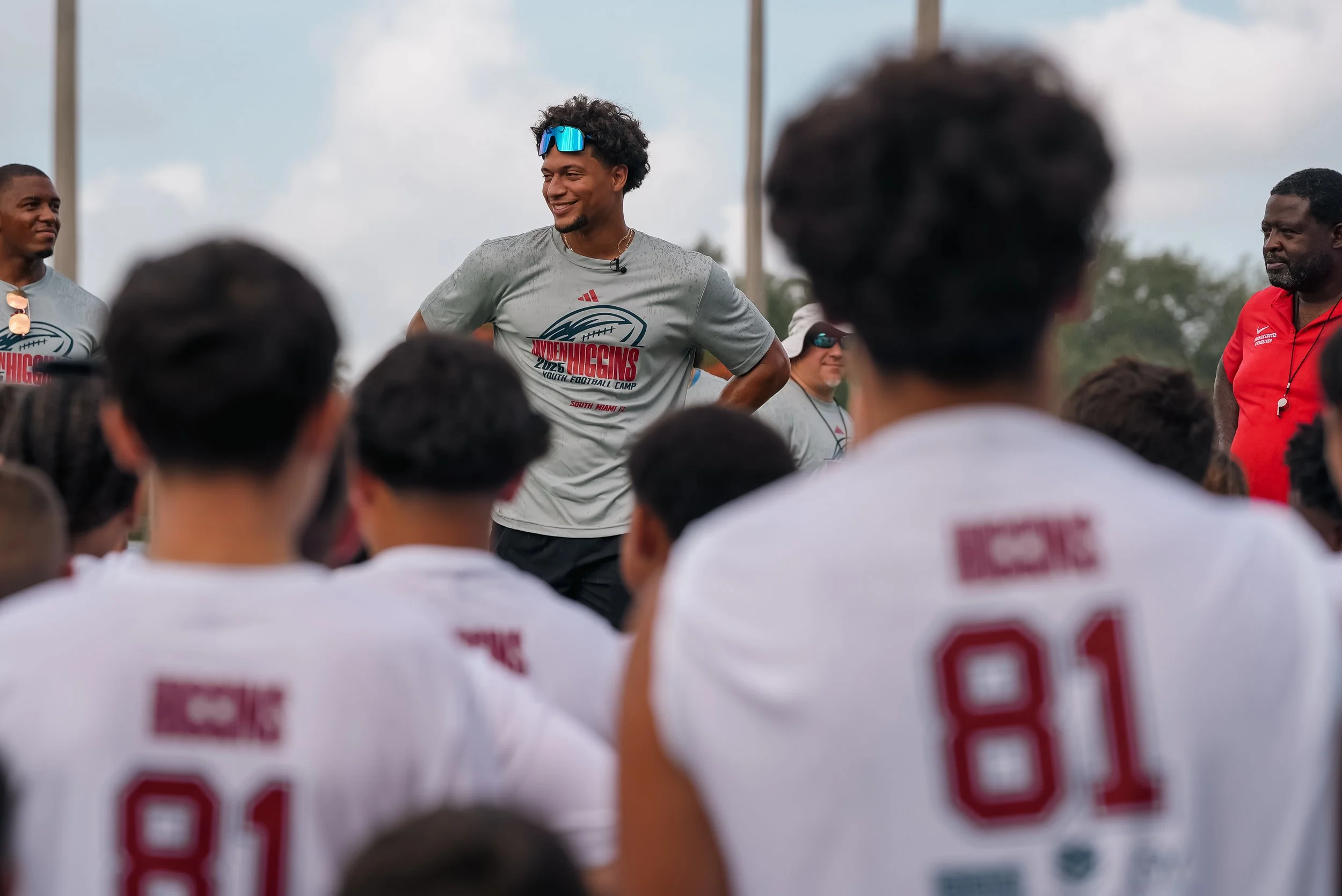Youth football camp with young athletes wearing white jerseys and a man with sunglasses and a gray shirt speaking to the group.