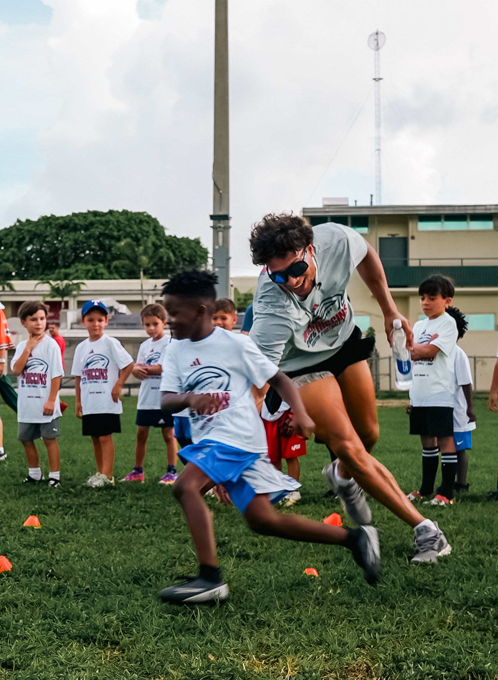 A young man wearing sunglasses and a white T-shirt participates in a race with children on a grassy field, surrounded by other kids in white T-shirts, with an overcast sky and a building in the background.