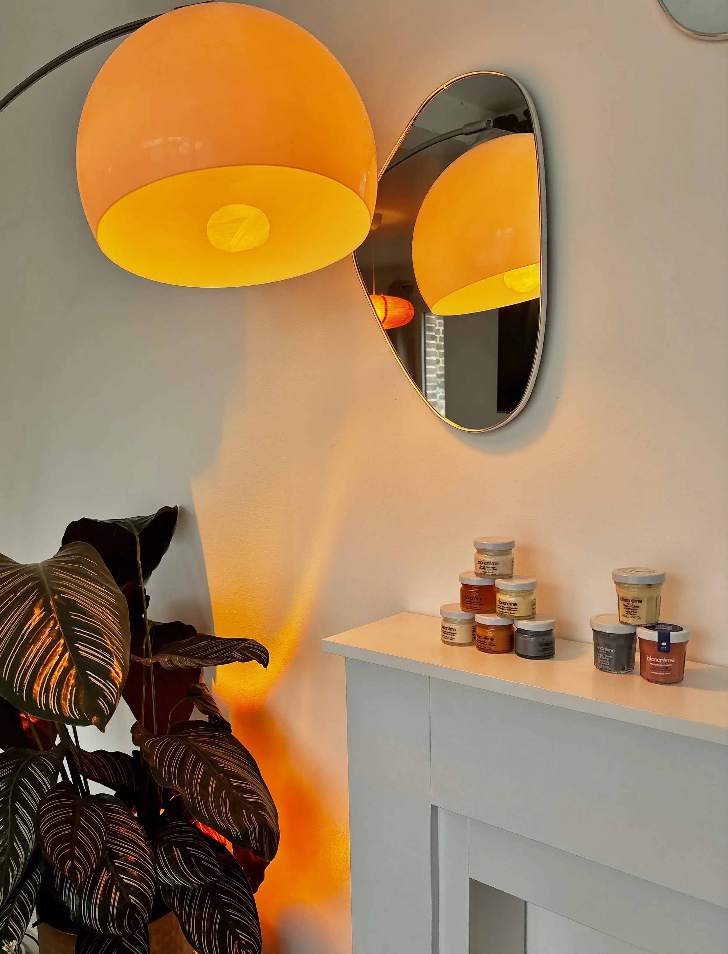 Une lampe orange suspendue au plafond, un miroir rond fixé au mur, une grande feuille de plante verte et une pile de pots de crème sur une table blanche dans une pièce bien éclairée.