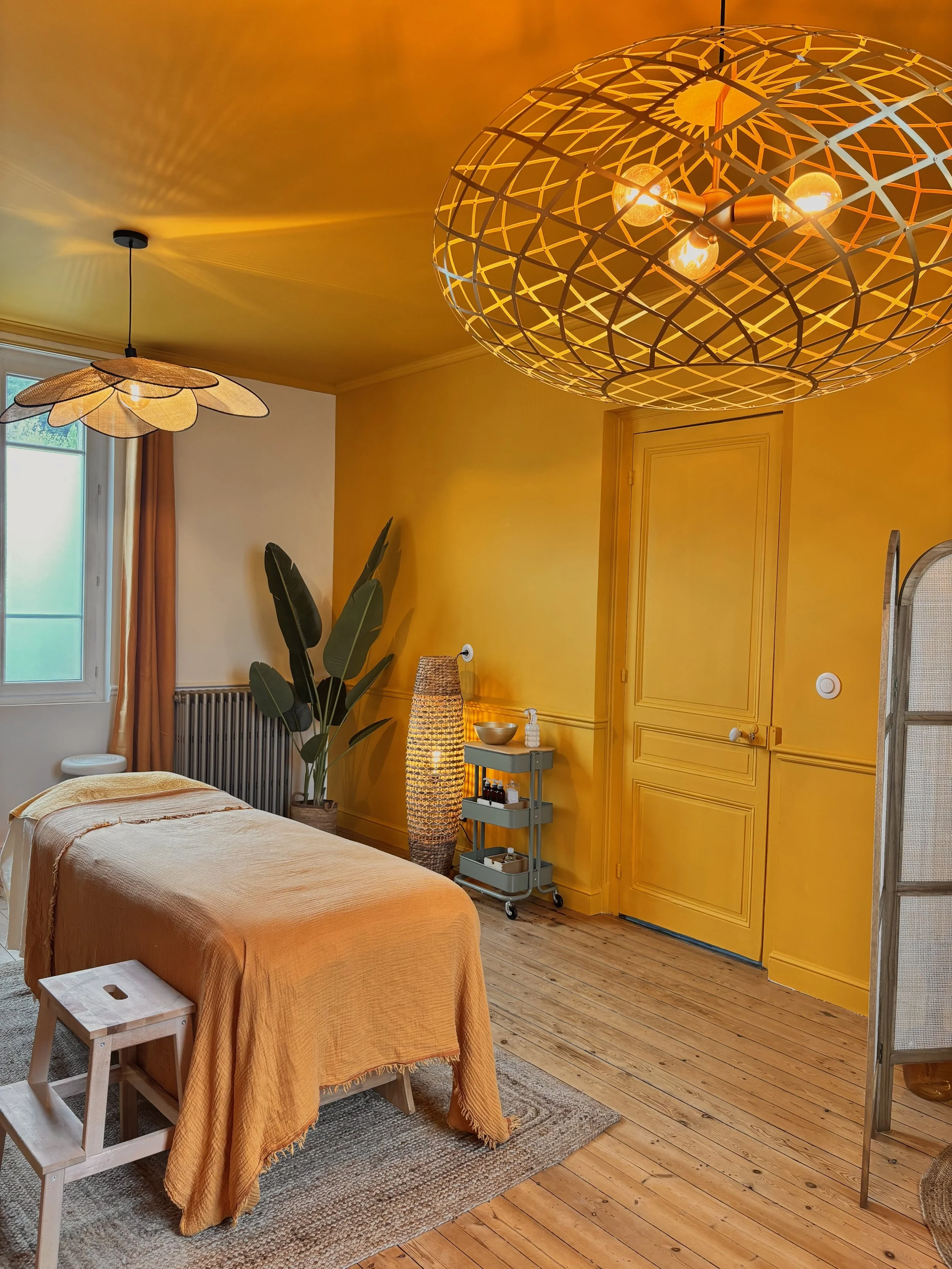 Chambre à coucher avec un lit, une lampe en osier, une plante verte, une armoire jaune, et deux lampes suspendues, une près de la fenêtre et l'autre au plafond.