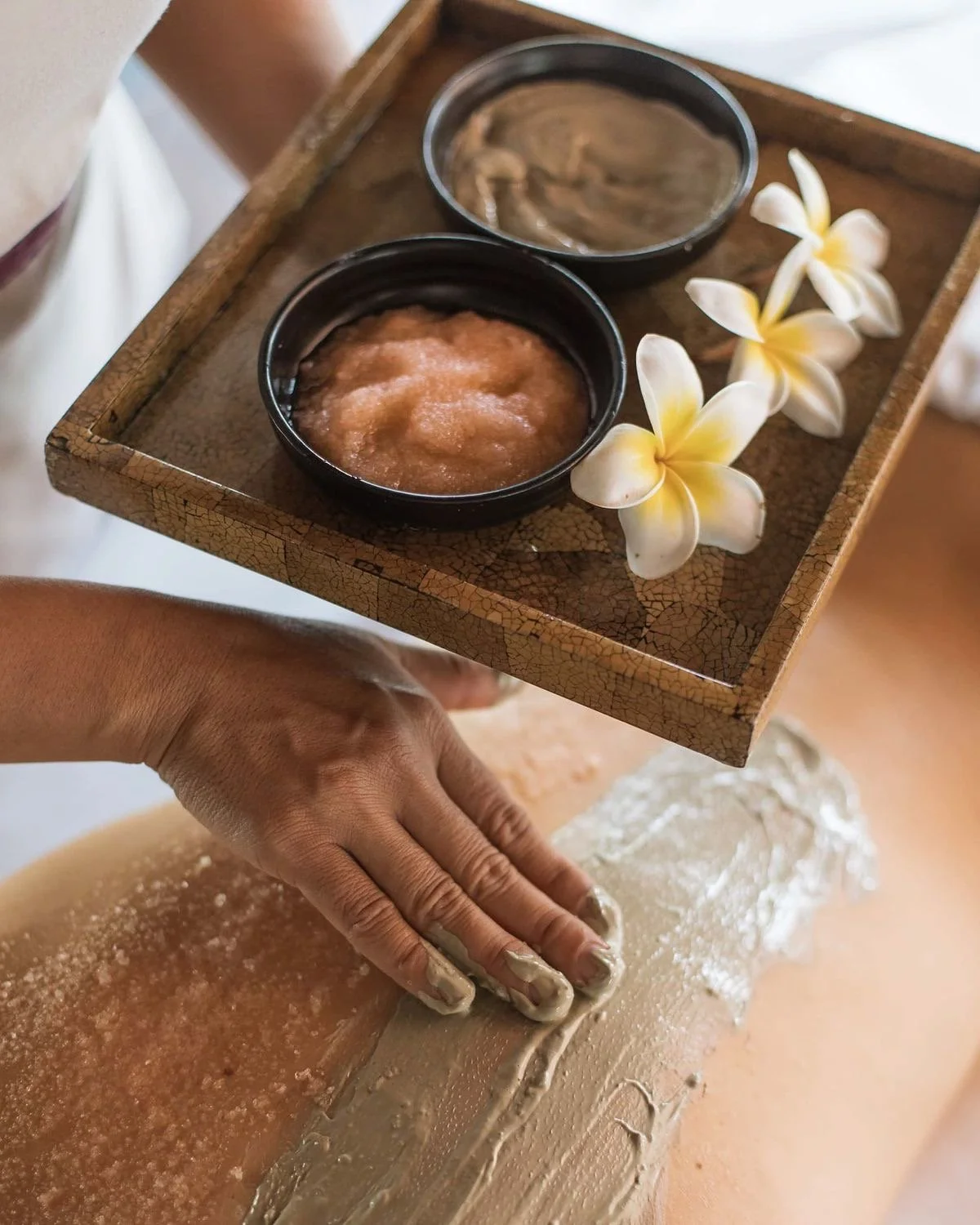 Une personne recevant un massage corporel dans un spa, avec un plateau en bois contenant deux petits bols avec des produits de soin et des fleurs blanches et jaunes.