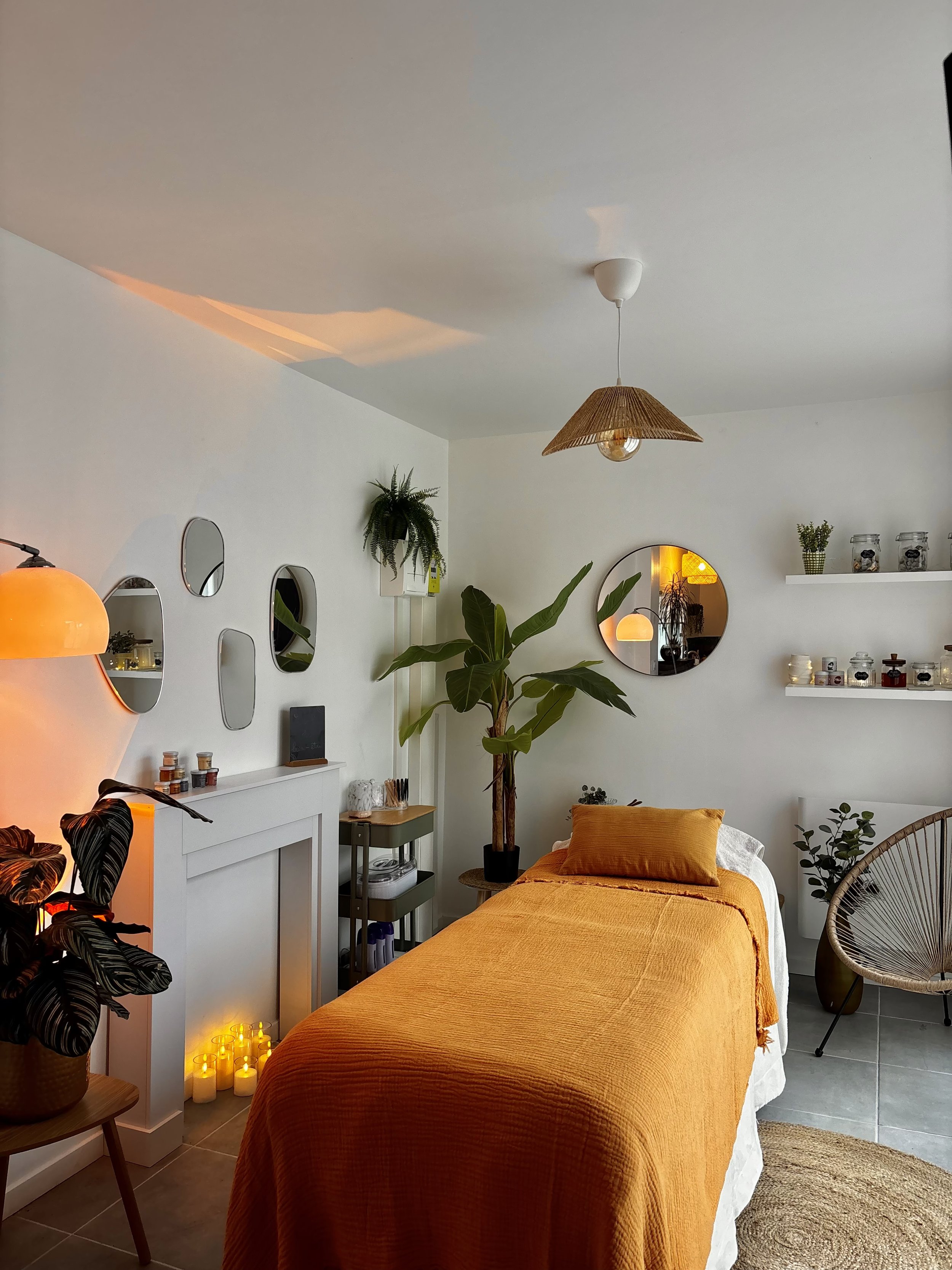 Une chambre décorée avec un lit recouvert d'une housse orange, entourée de plantes d'intérieur, de miroirs, d'ampoules suspendues, et de plusieurs étagères avec des bocaux et des décorations. Un lampadaire dégage une lumière chaleureuse et il y a un tapis en rotin au sol.