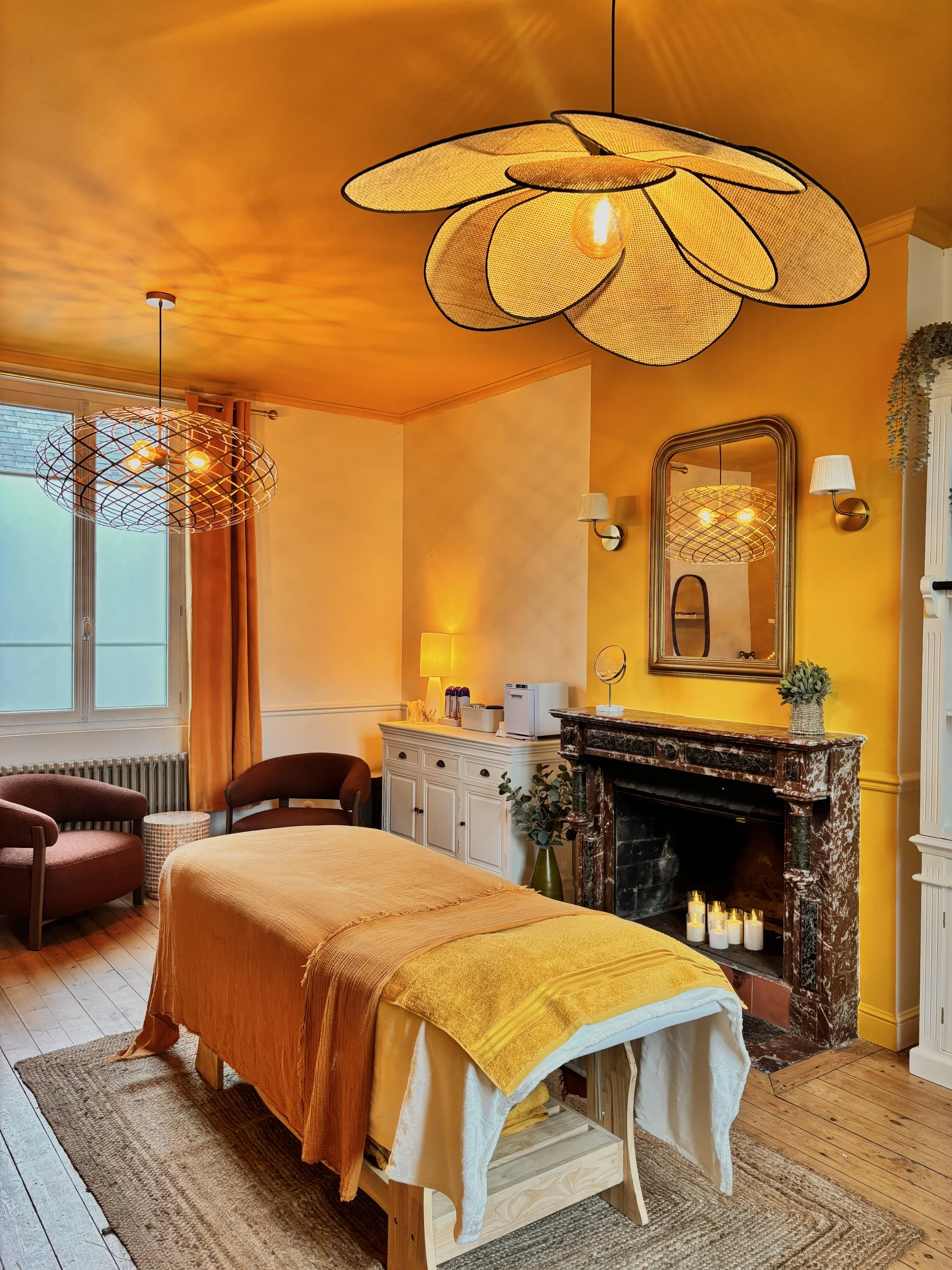Une chambre avec un lit, une cheminée en marbre noir avec bougies, un miroir, deux appliques murales et des lampes de chevet, décorée en jaune et orange.