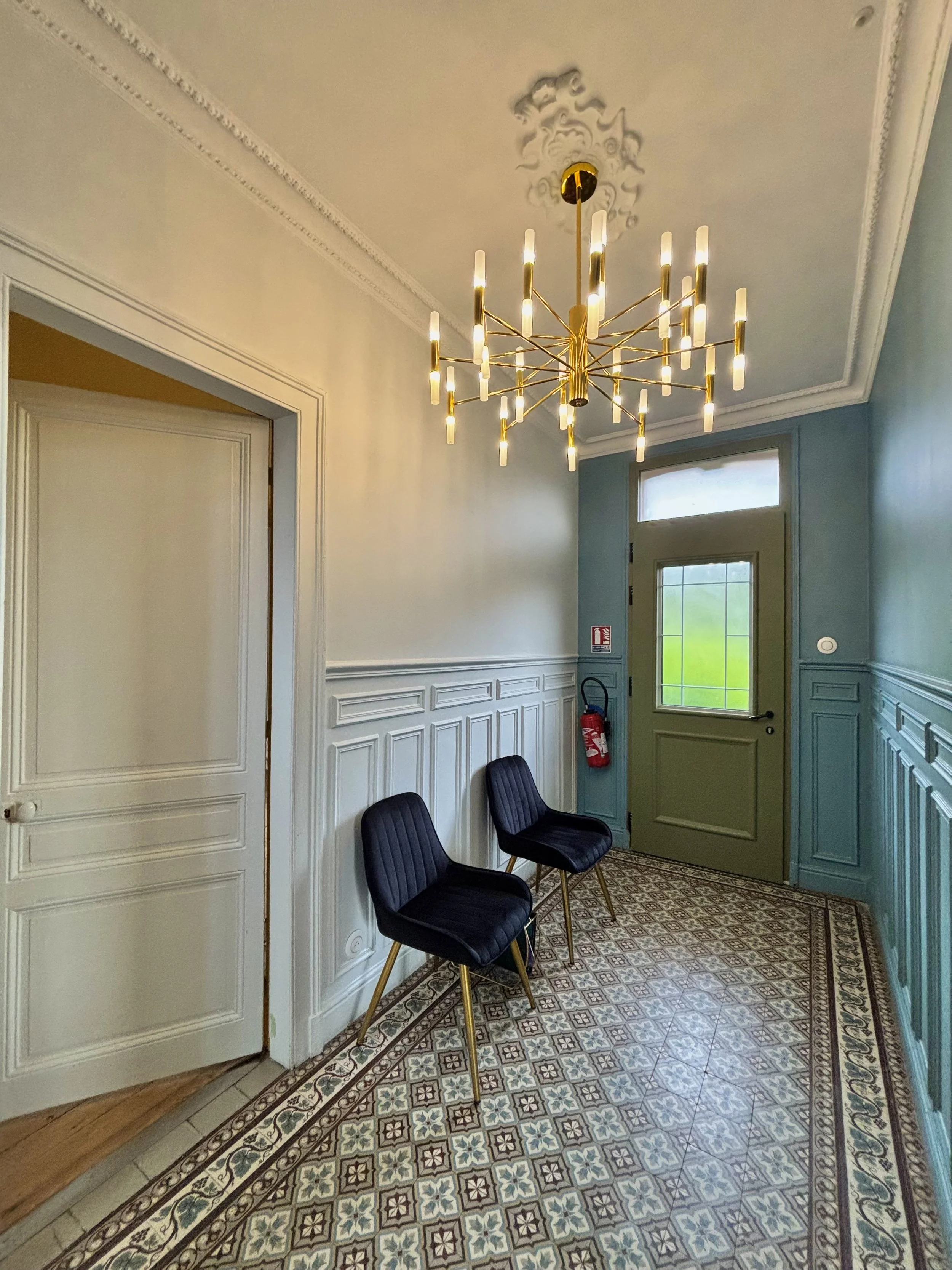 Une salle d'attente avec deux chaises noires, une porte verte, une fenêtre, un tapis aux motifs géométriques, un lustre doré suspendu au plafond, et un détecteur de fumée sur le mur.