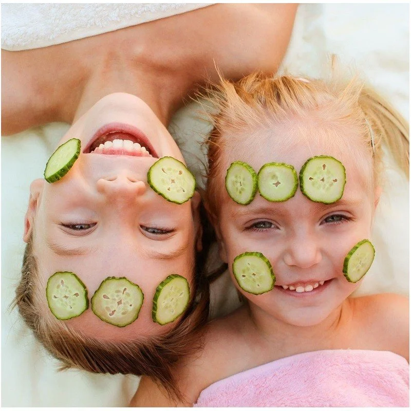 Deux enfants avec des tranches de concombre sur le visage, souriant et allongés sur un lit, créant une scène amusante et relaxante.