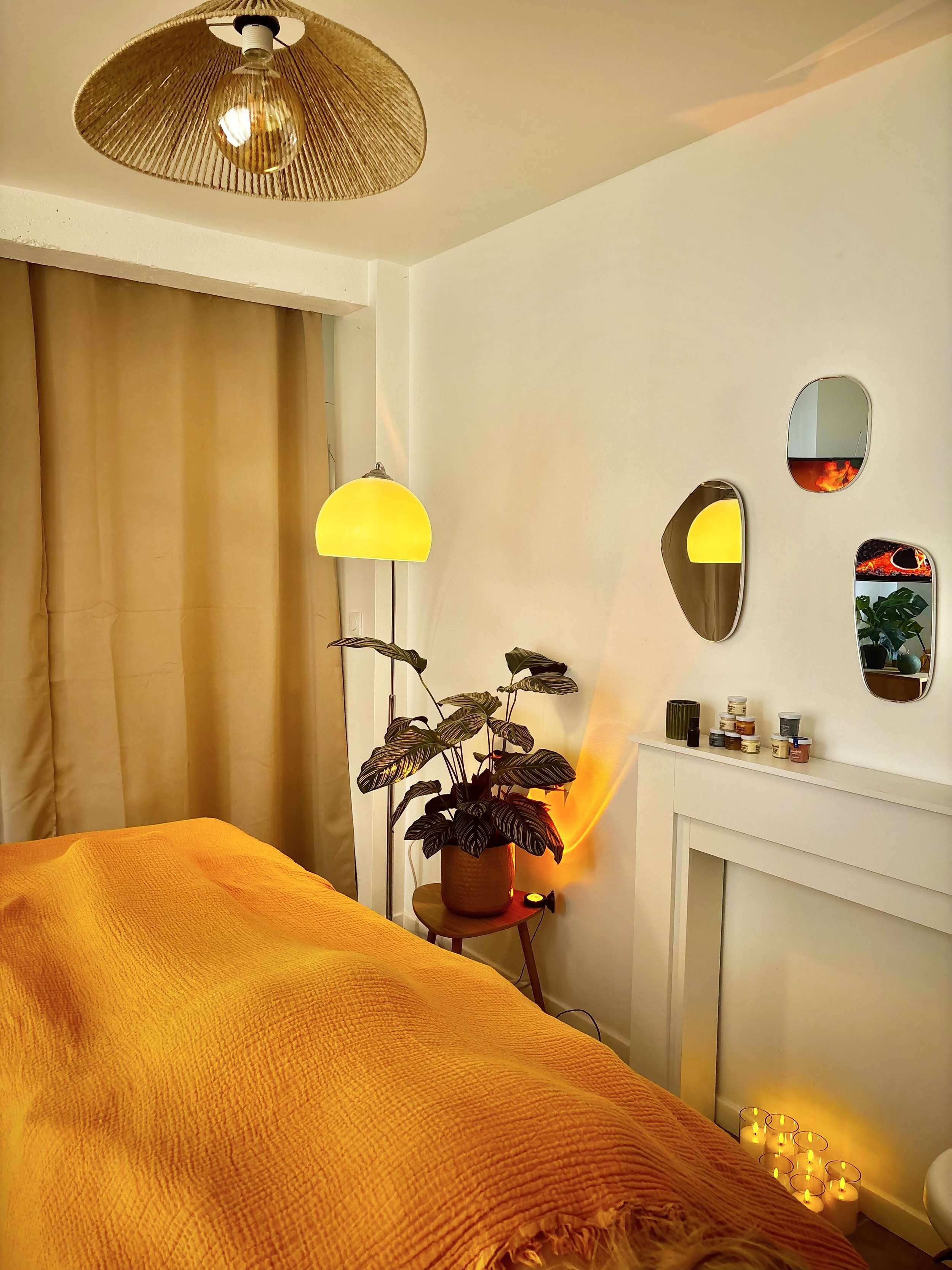 Une chambre chaleureuse avec un lit recouvert d'une couverture orange, une plante dans un pot en osier, une lampe à pied avec un abat-jour jaune, un lampadaire avec un abat-jour en rotin et une ampoule suspendue, un mur blanc décoré de miroirs et une petite étagère avec des pots de plantes et des objets, et des bougies allumées sur le sol.