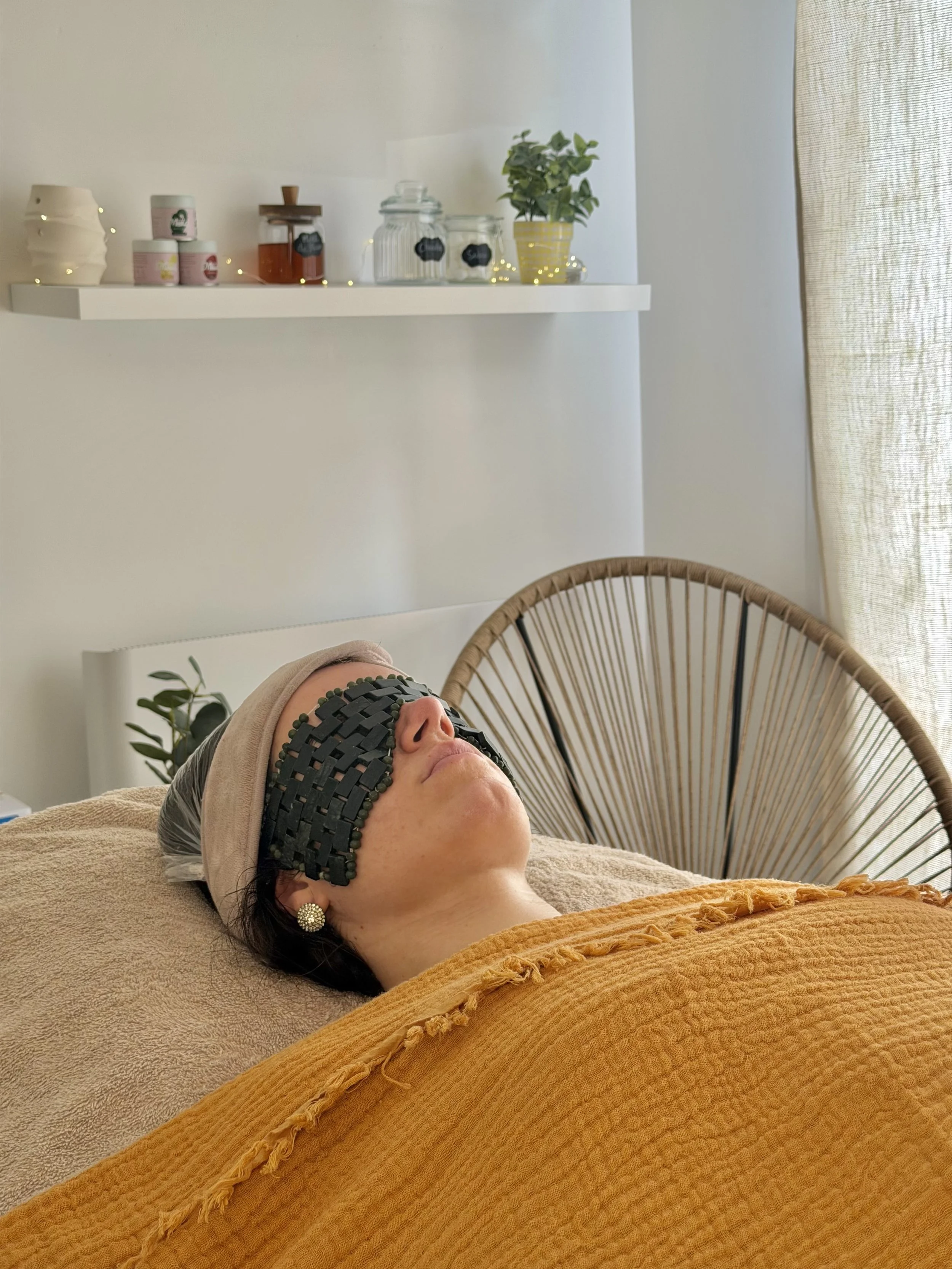 Une femme repose sur une table de spa avec un masque en pierre noire sur les yeux, dans une ambiance relaxante avec des plantes et des bibelots sur une étagère en arrière-plan.