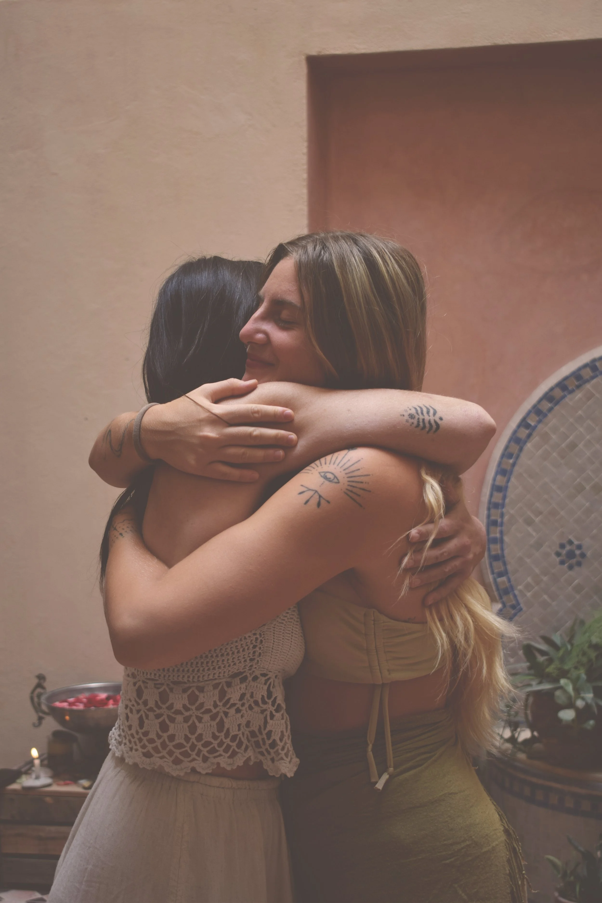Two women hugging each other warmly in a cozy indoor setting, both smiling with eyes closed.