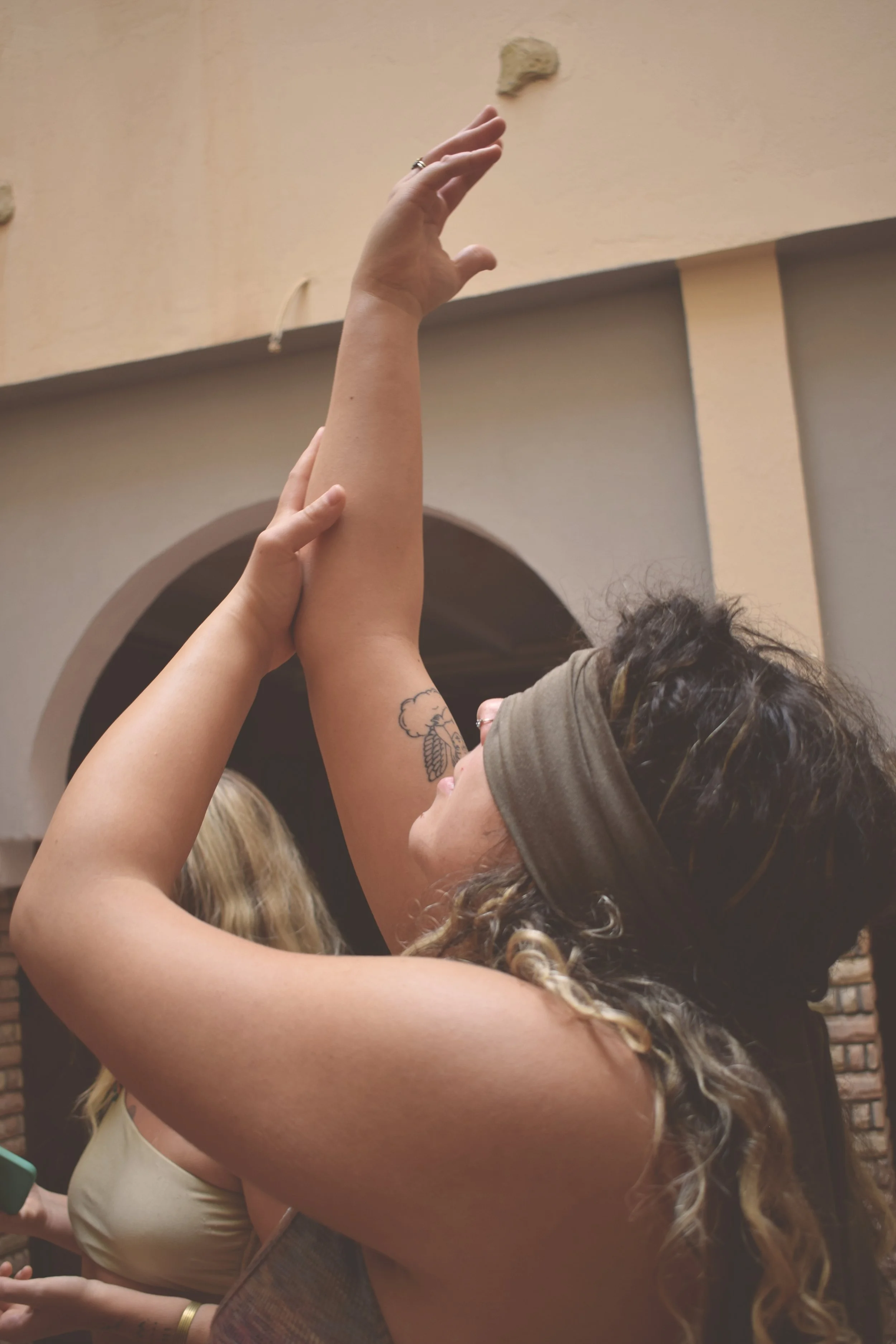 A woman with curly hair wearing a headband and tank top, stretching her arm upward while another person helps support her elbow. The background shows a building with an arched doorway and beige walls.
