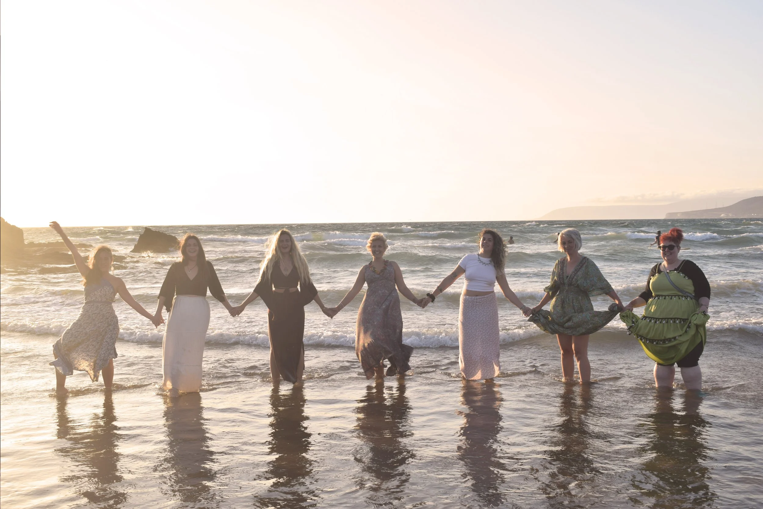 Seven women standing in the ocean holding hands during sunset, dressed in colorful dresses.