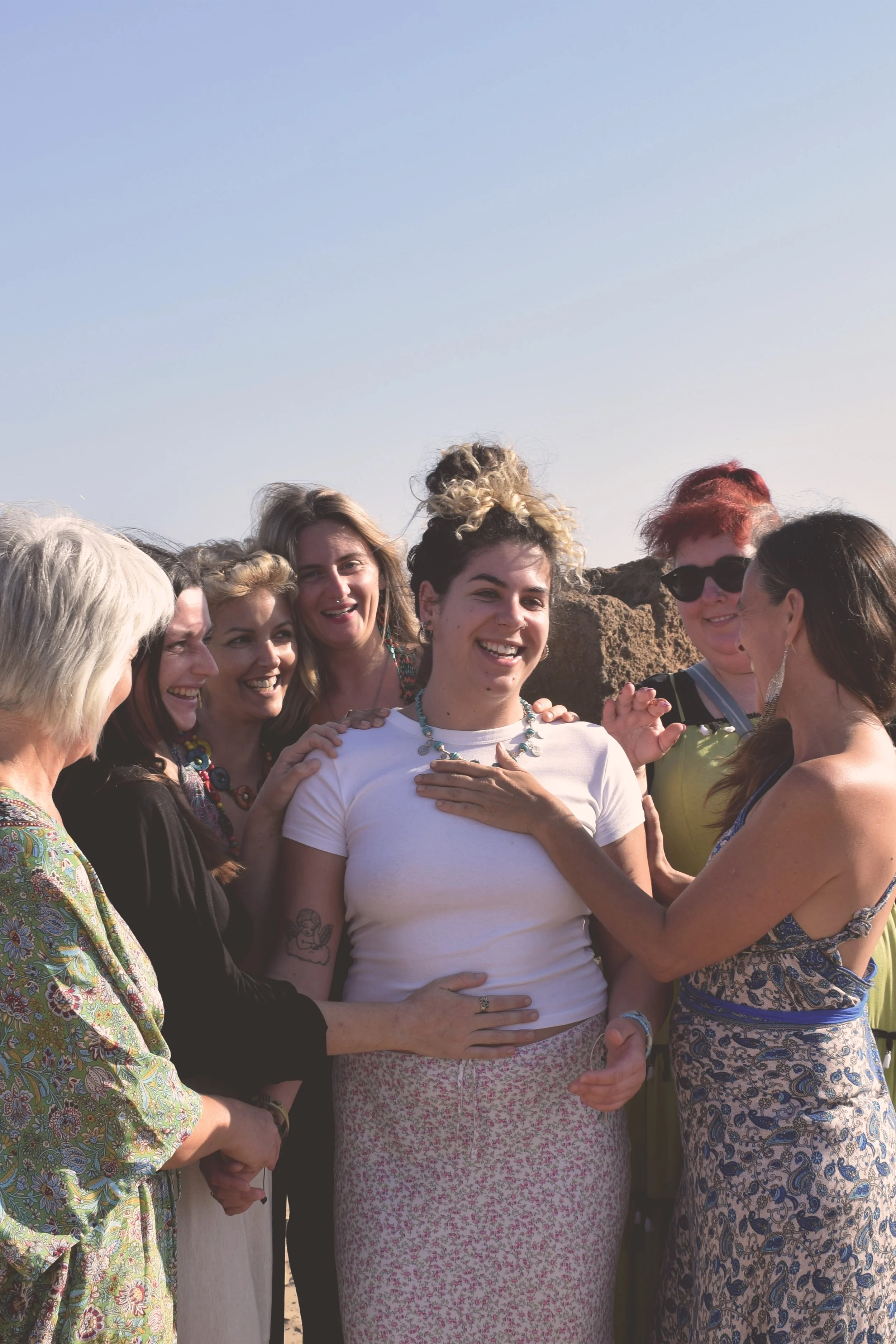 A group of women gathered together outdoors, smiling and touching a woman in a white shirt who is holding her hand over her heart. The group appears to be happy and engaged in a moment of celebration.