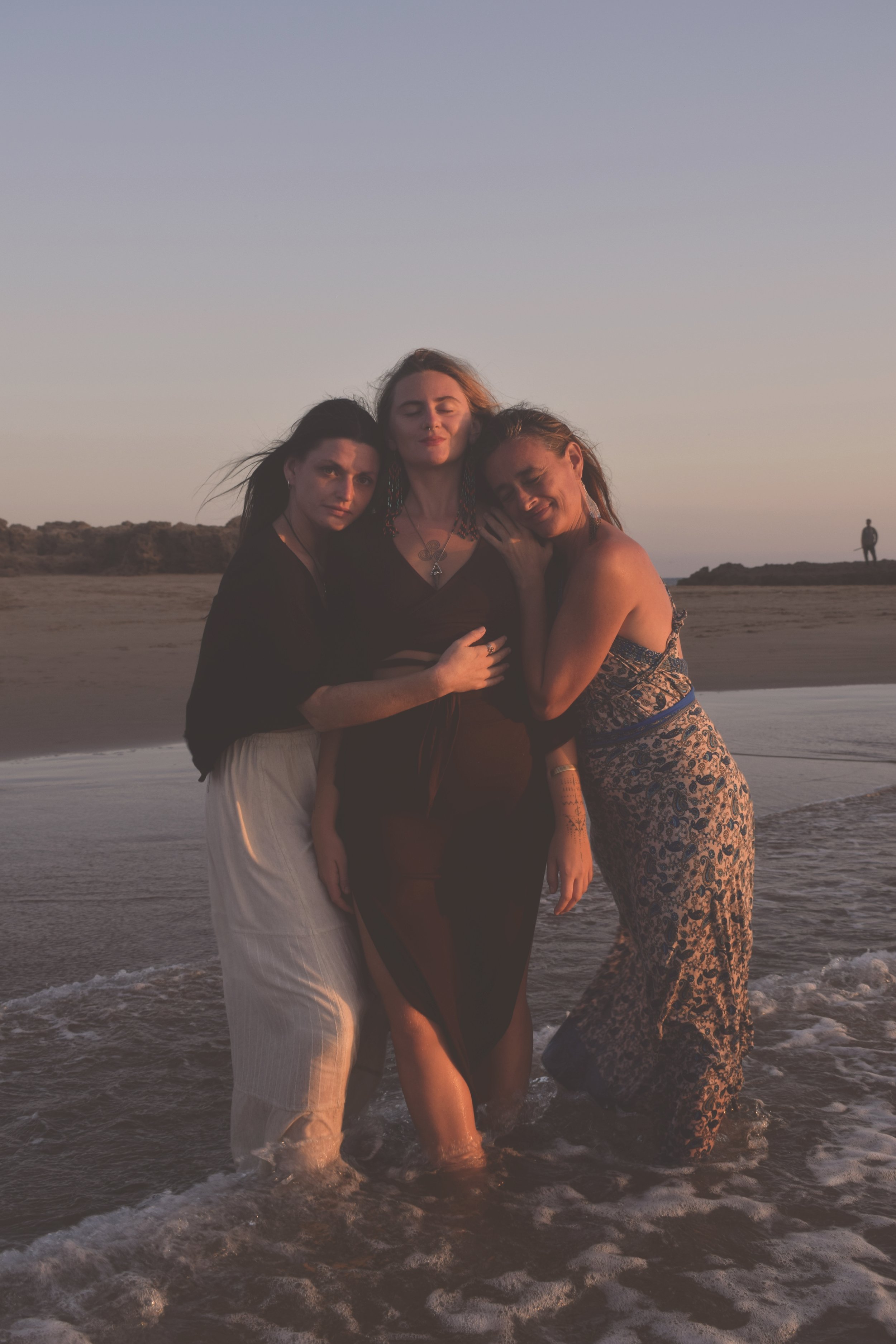 Three women standing in the ocean at sunset, embracing each other and smiling, with a person walking on the beach in the background.