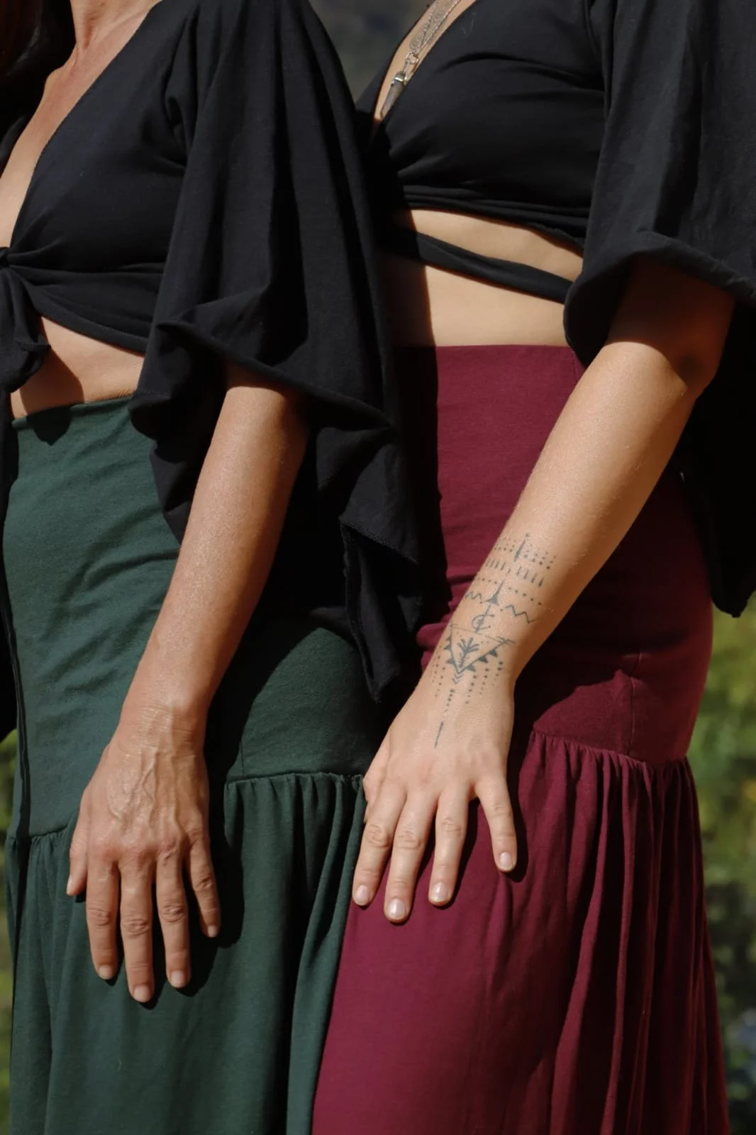 Close-up of two women standing side by side outdoors, showing their torsos and hands. One wears a black top with green skirt, the other a black top with maroon skirt. The woman in maroon has a tattoo on her forearm.
