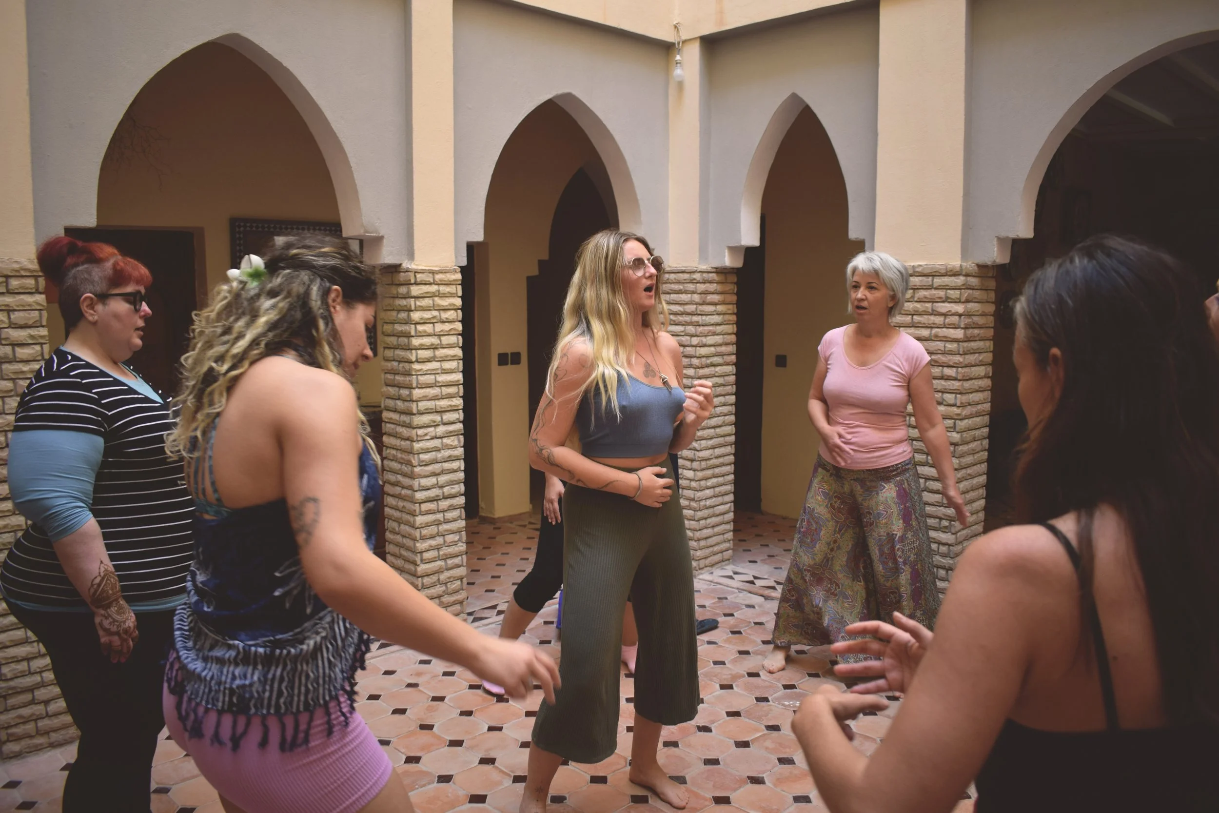 Group of women dancing and socializing in an indoor courtyard with brick archways and tile floor.