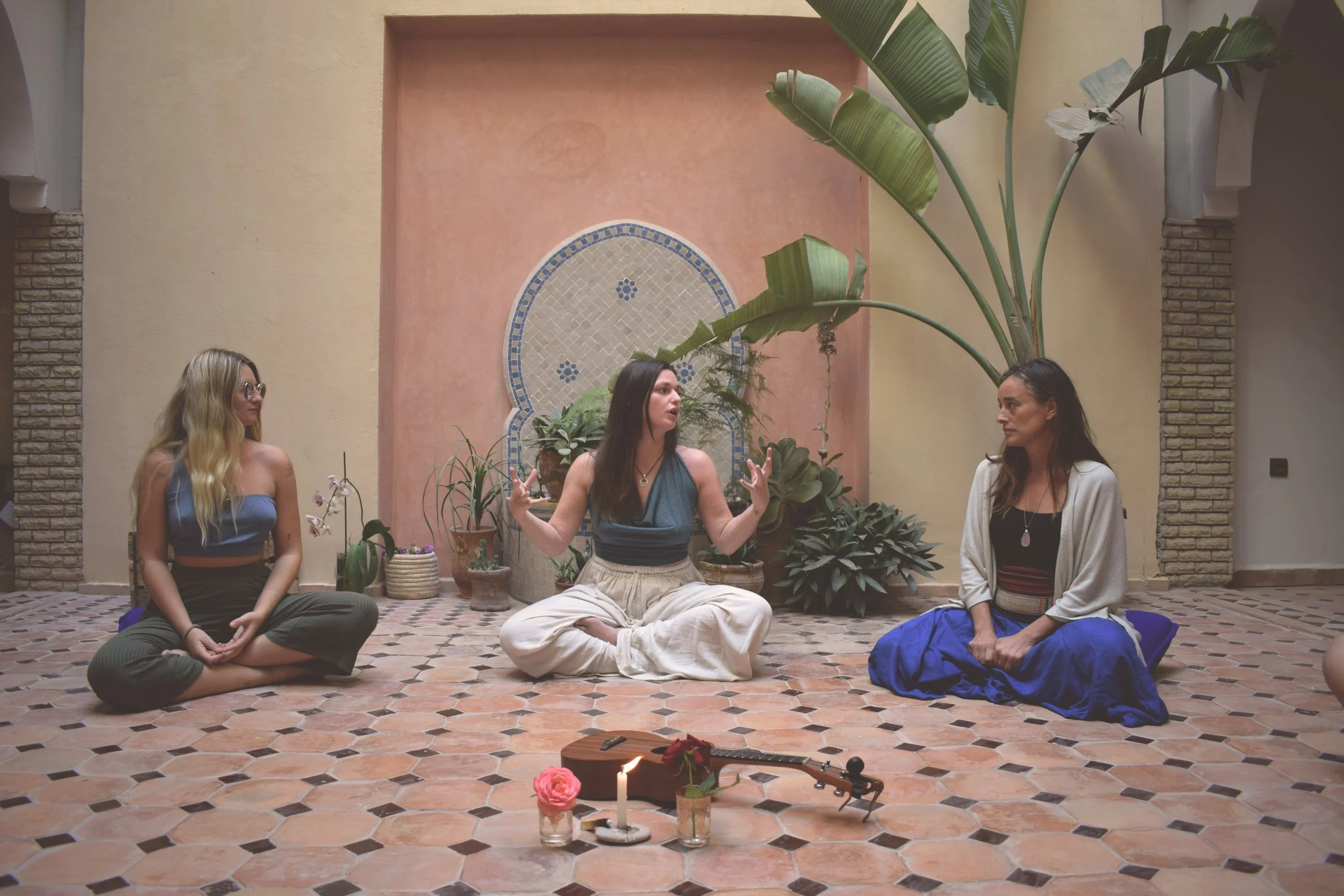 Three women sitting cross-legged on a tiled floor during a spiritual or meditation session, with candles, a journal, a guitar, pink roses, and green plants in a decorative indoor setting.