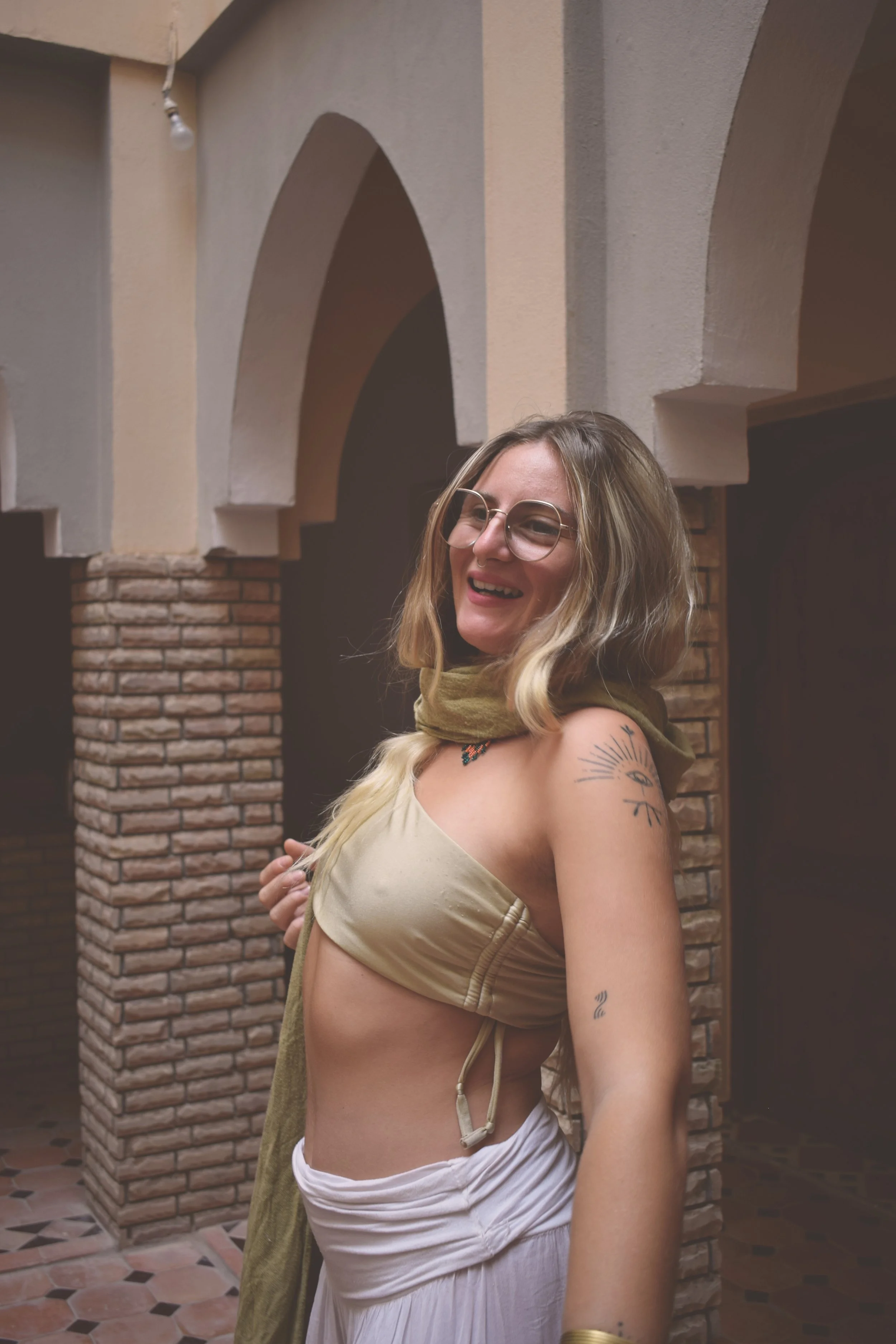 A smiling woman with glasses and tattoos wearing a beige top and white skirt, standing indoors near brick arches.
