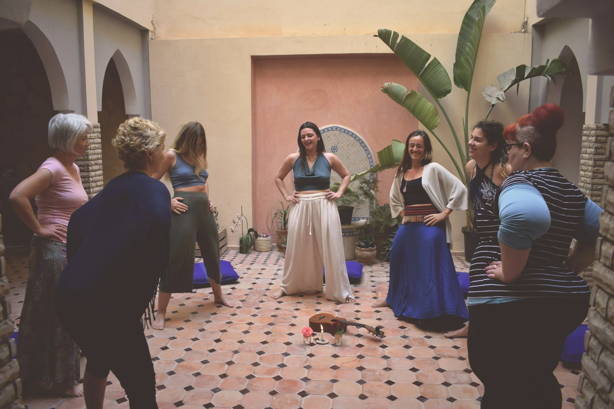 Group of women standing in a circle indoors, smiling and engaging in a group activity around a small altar with candles, flowers, and a guitar, in a room with tropical plants and patterned floor tiles.