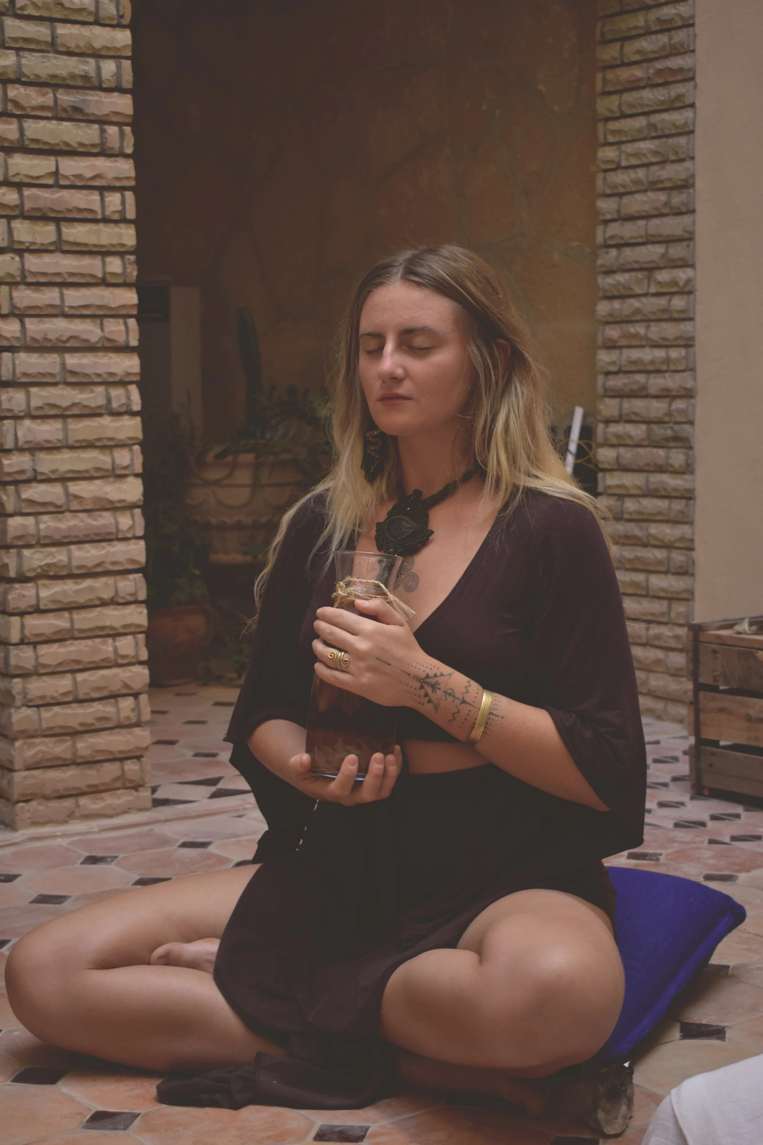 A woman sitting cross-legged on a tiled floor with eyes closed, holding a glass container with a plant, in a cozy room with brick and stone walls, dressed in dark clothing and wearing accessories.