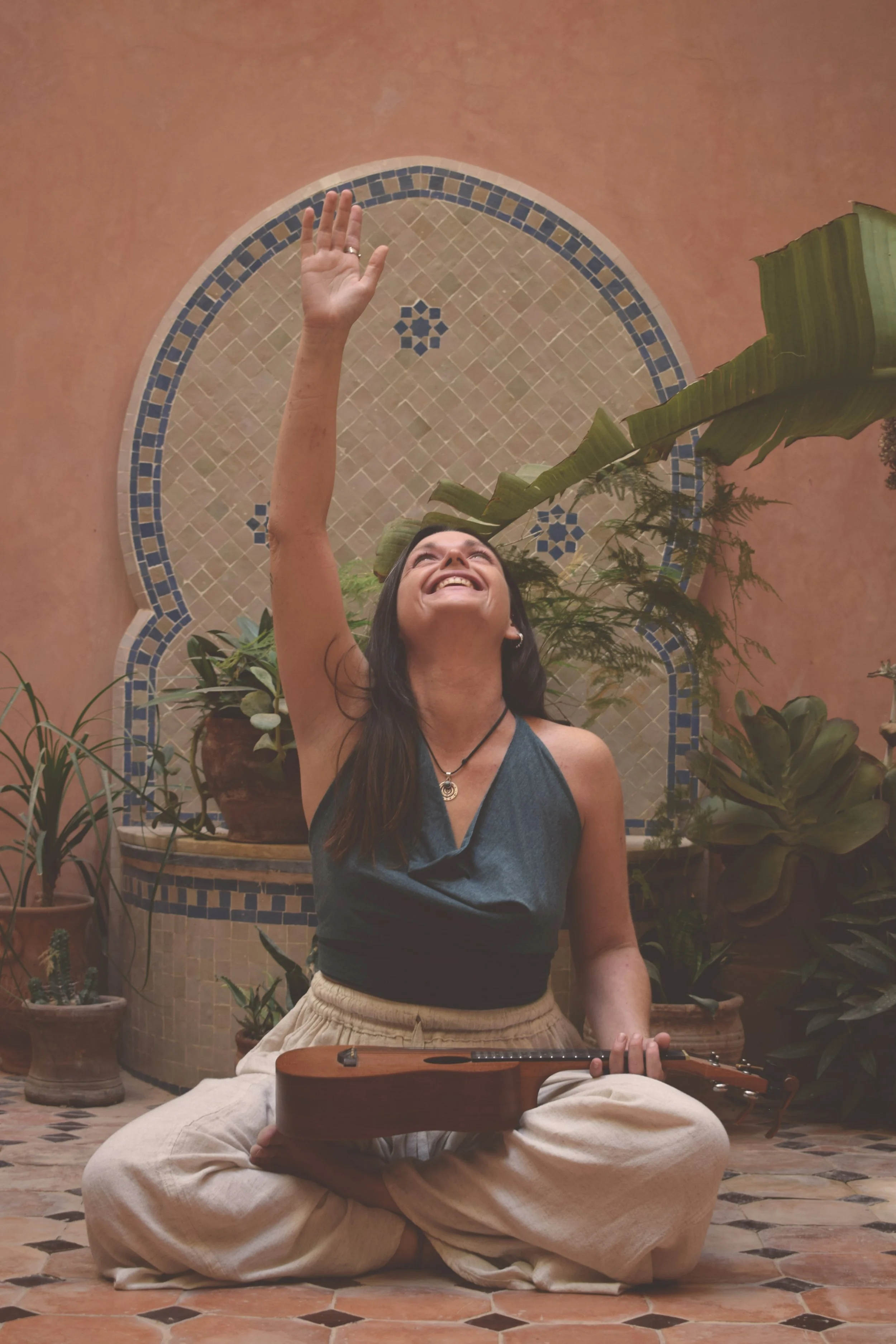 A woman sitting cross-legged on a tiled floor, playing guitar, raising her right arm, smiling and looking up, surrounded by potted plants, with an arched mosaic wall behind her.