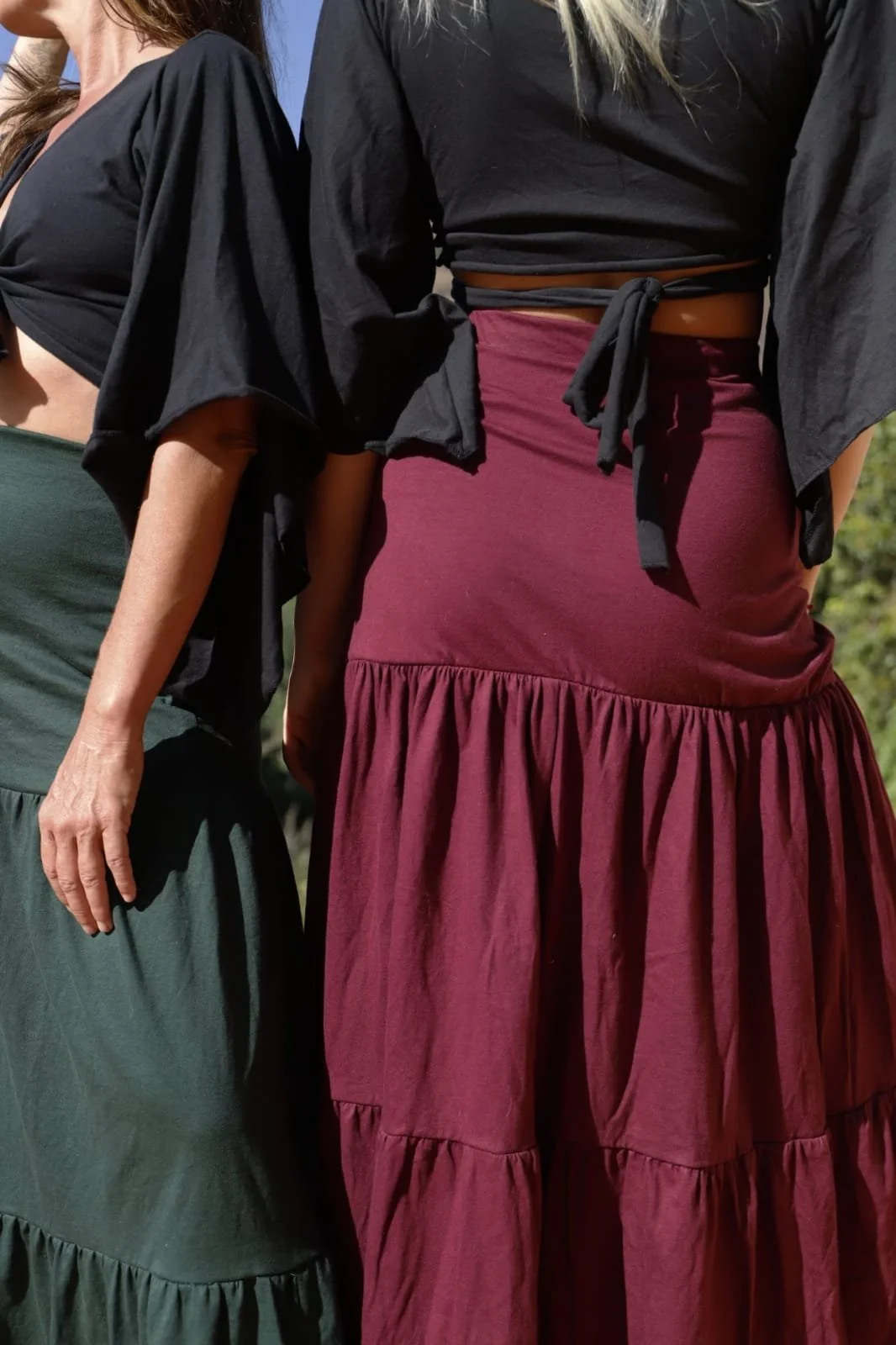 Close-up of two women standing side by side outdoors. One woman has a green skirt and a black top with puffy sleeves. The other woman has a maroon skirt with ruffles, a black top with a tied front, and a tan belt.