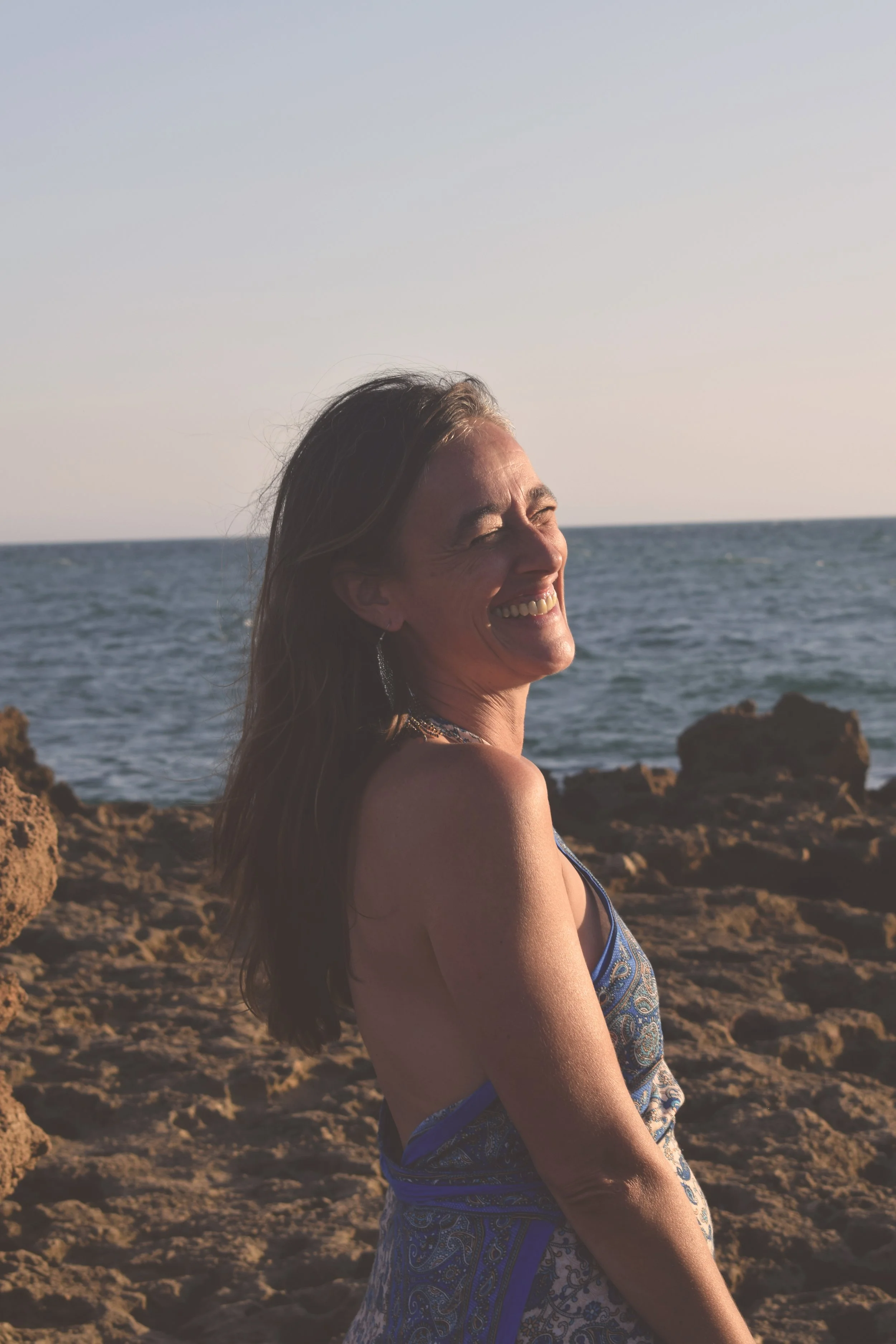 A woman smiling at the beach during sunset, with ocean waves and rocky shoreline in the background.