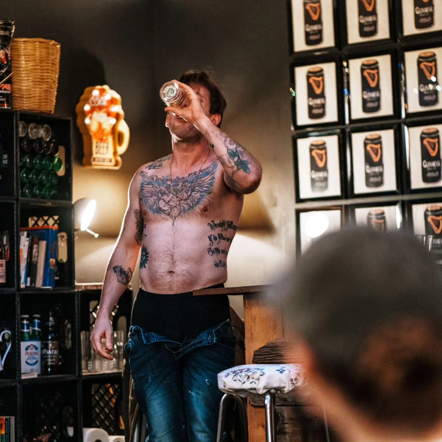 A shirtless man with tattoos drinking from a glass in a dimly lit bar or pub, with shelves of bottles and framed posters on the wall.