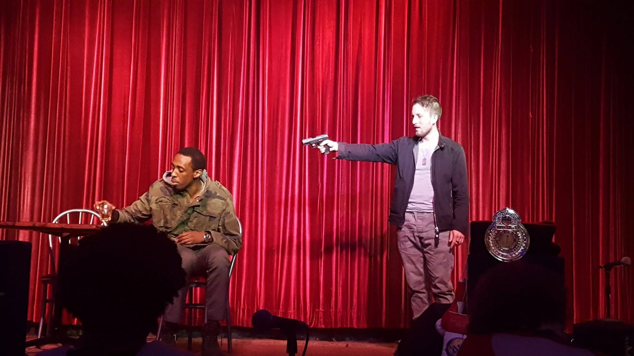 Two men on stage in front of a red curtain; one sitting at a table holding a glass, the other standing and pointing a gun at the seated man.