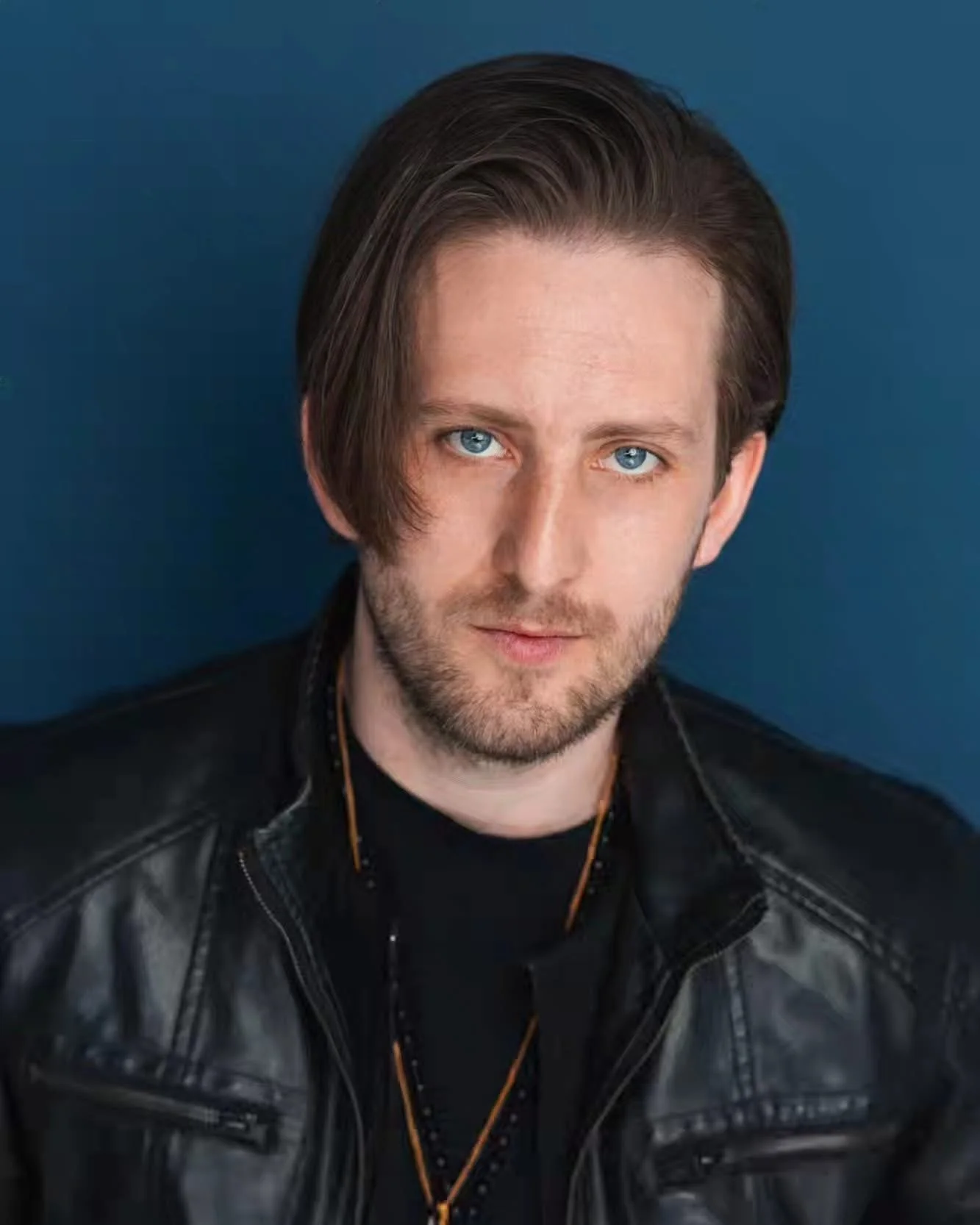 Portrait of Michael Joseph Delaney, a man with medium-length brown hair and blue eyes, wearing a black leather jacket against a blue background.