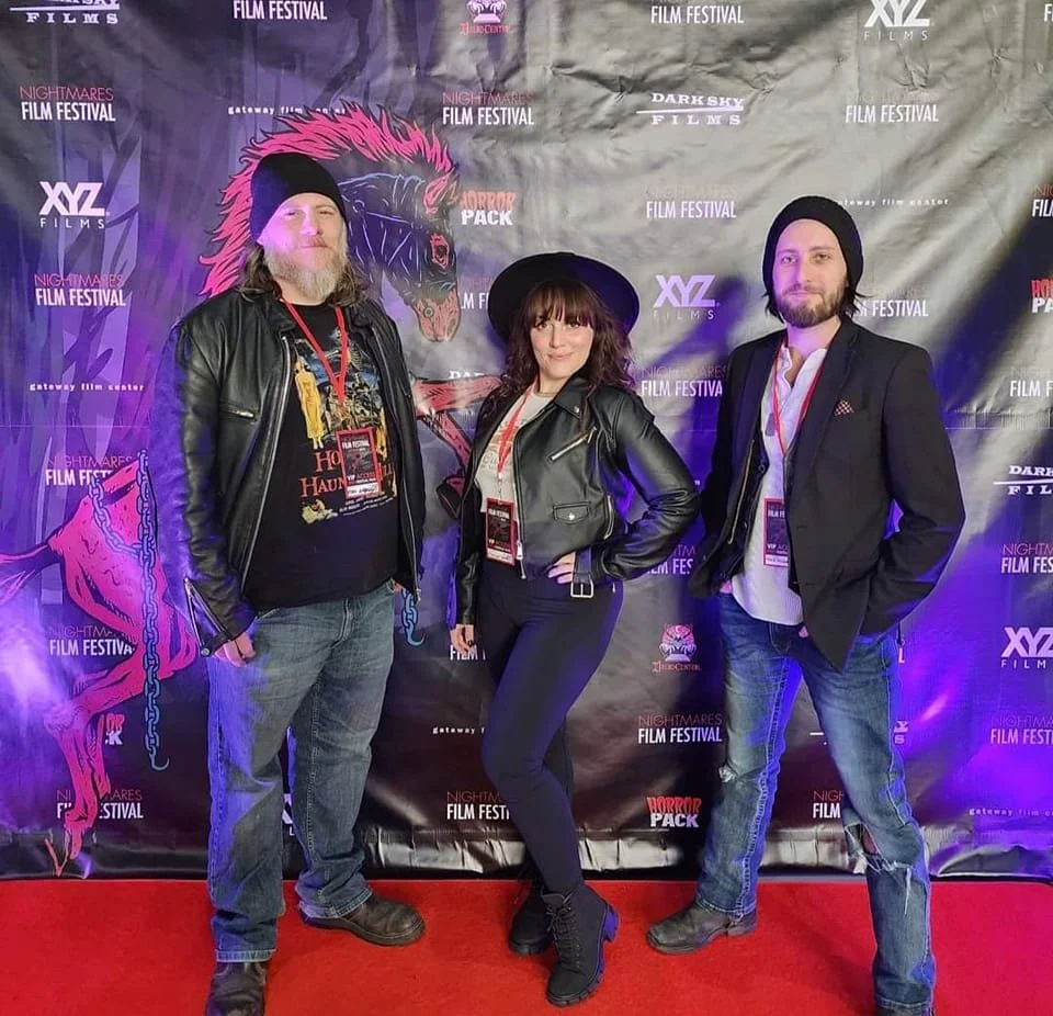 Three people standing on a red carpet at a film festival, posing in front of a backdrop with horror-themed graphics and logos.