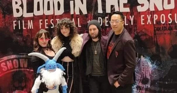 Four people and a puppet of a blue creature with horns in front of a Blood in the Snow film festival banner.