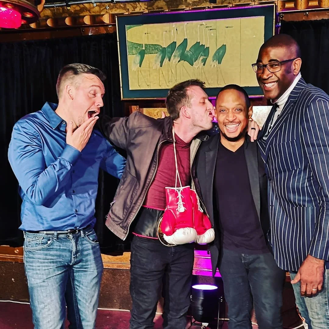 Four men posing together in a bar or club, with one man wearing boxing gloves hanging around his neck. They are smiling and joking, with colorful lights and an abstract painting in the background.