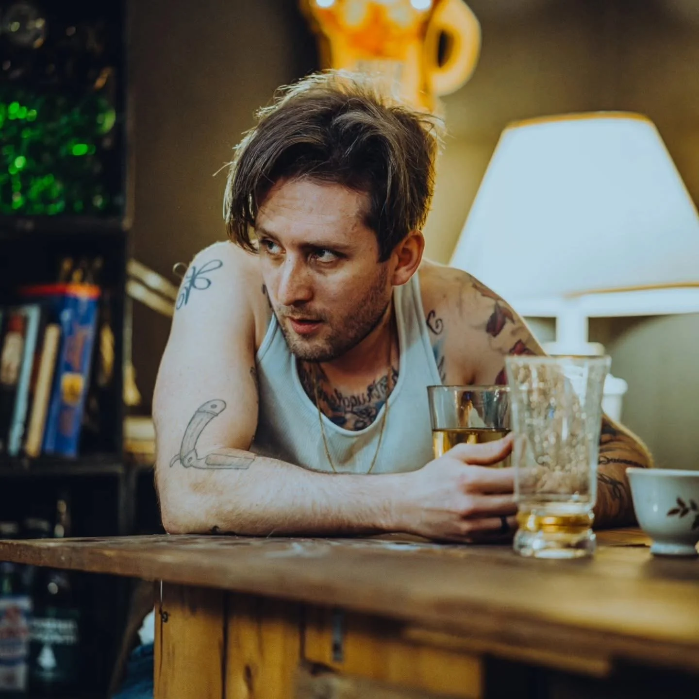 A young man with tattoos, wearing a white tank top, sitting at a wooden table with drinks, in a cozy indoor setting with a lamp and bookshelf in the background.