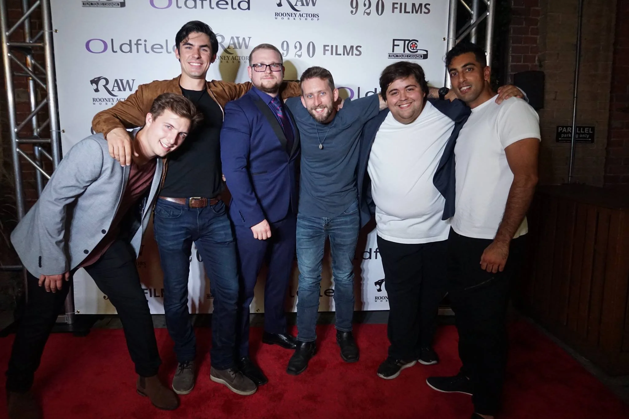 Group of seven men smiling and posing together in front of a backdrop with logos, on a red carpet at an event.