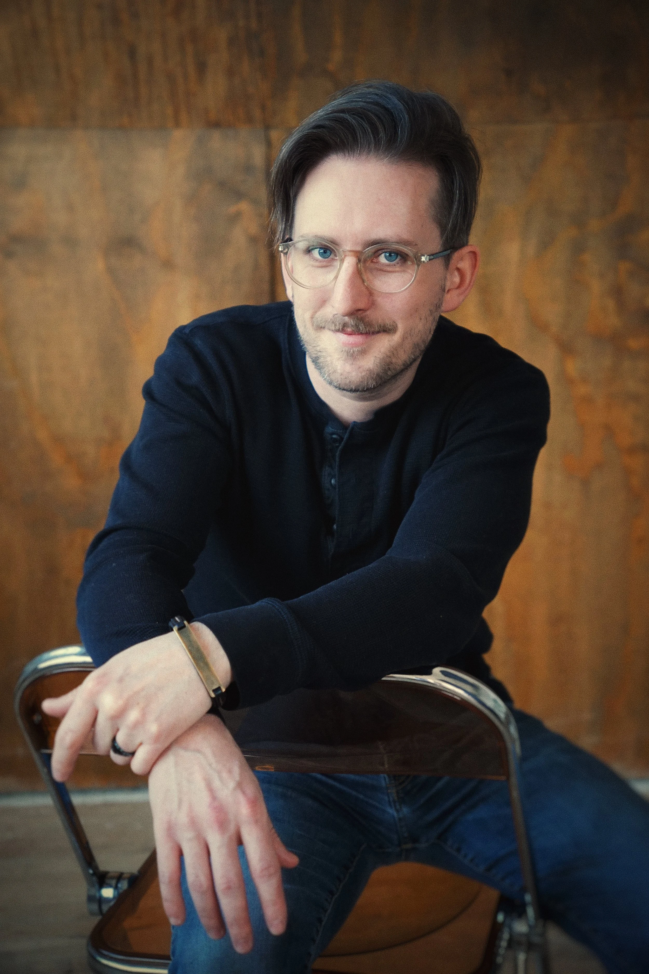 Michael Joseph Delaney, a man with glasses and a slight smile, wearing a black shirt and blue jeans, sitting on a chair with a wooden back, in front of a wooden wall.
