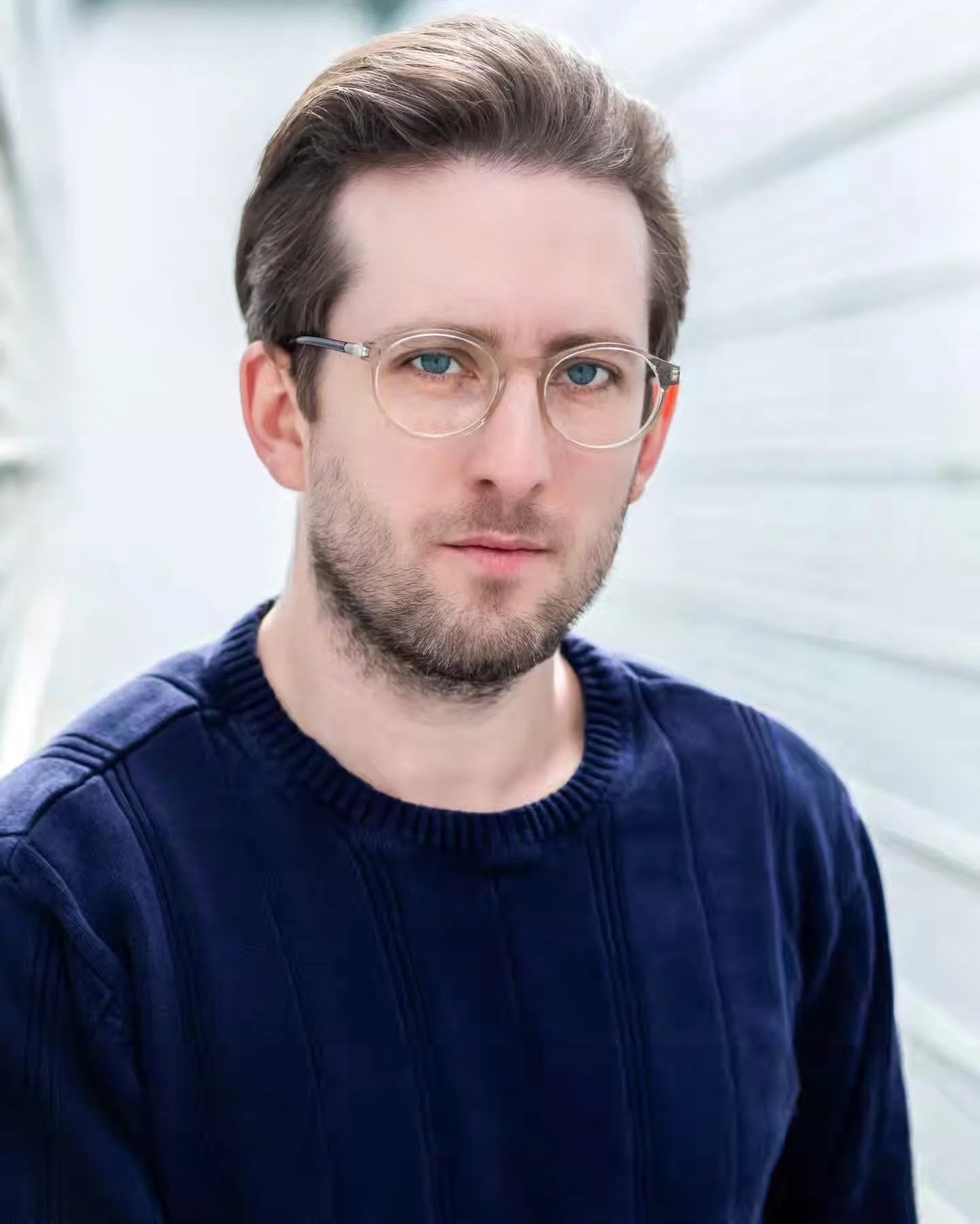 A young man (Michael Joseph Delaney) with brown hair, glasses, and a beard, wearing a navy blue sweater, standing in a bright, white, outdoor or greenhouse setting.