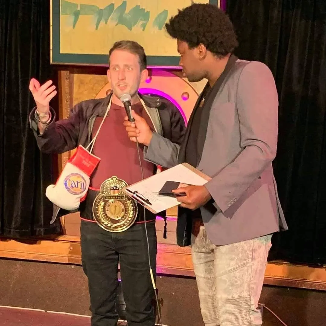 Two men are standing on a stage, one holding a microphone and a large championship belt around his waist. The man with the belt is speaking, while the other man is holding a clipboard and listening. The background features black curtains and framed artwork.