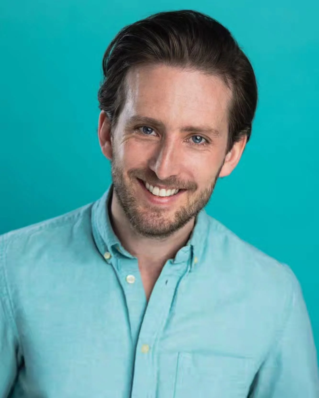 Michael Joseph Delaney, man with short brown hair, blue eyes, and a beard, smiling at the camera, wearing a light blue button-up shirt against a teal background.