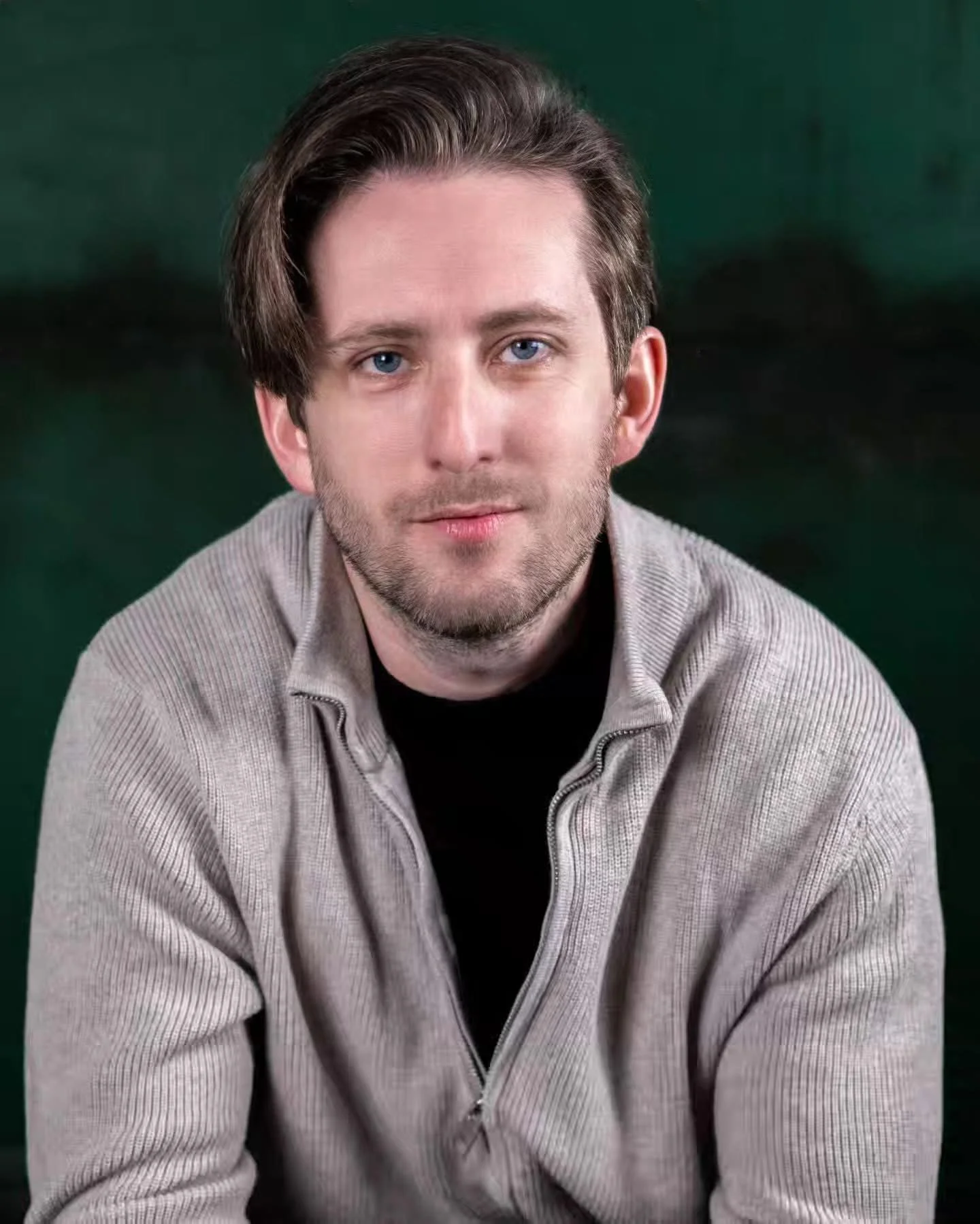 Portrait of a young man, Michael Joseph Delaney, with light brown hair, blue eyes, and a slight beard, wearing a light gray zip-up sweater over a black shirt, against a dark green background.