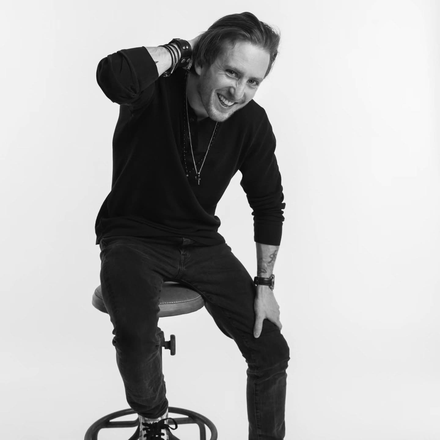 Michael Joseph Delaney, a young man with long hair sitting on a stool, smiling, wearing dark clothing, with his left hand on his thigh and right hand behind his head, against a plain white background.