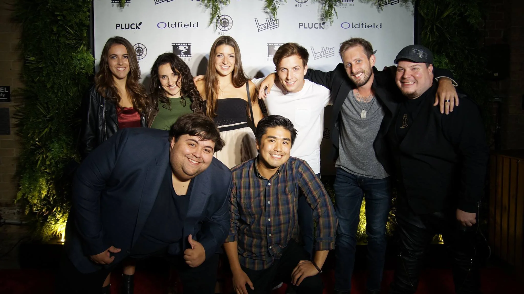Group of ten people posing and smiling at an event with a step and repeat backdrop featuring logos.