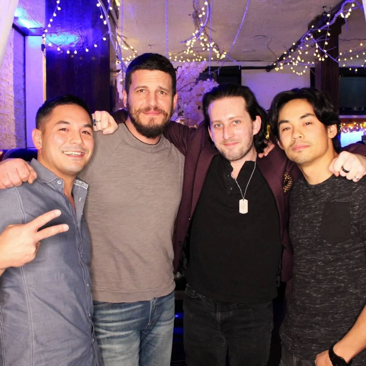 Four men posing together at a party with colorful string lights and a decorative tree in the background.