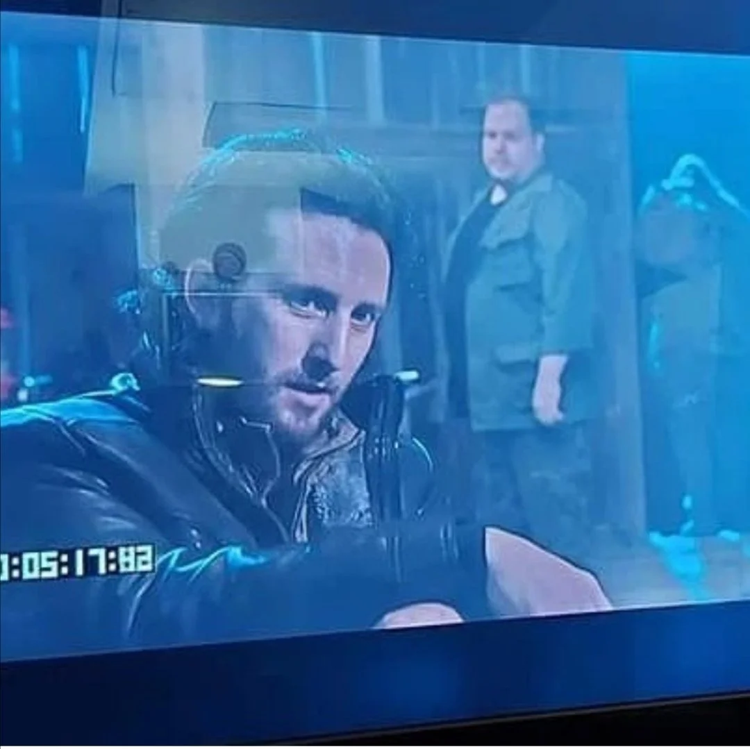 Scene from bts of a movie showing Michael Joseph Delaney, with long dark hair in the foreground, and another man standing in the background in a room with a coat rack.