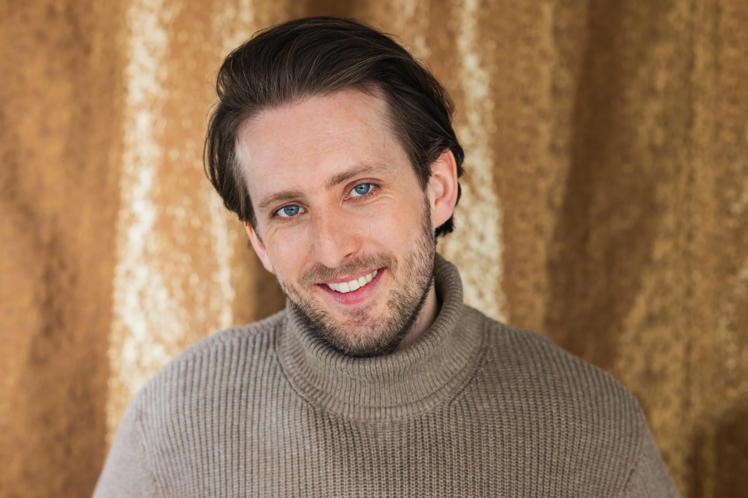 Portrait of a young man, Michael Joseph Delaney, with dark hair and blue eyes, smiling, wearing a beige turtleneck sweater, with a blurred brown and gold curtain background.