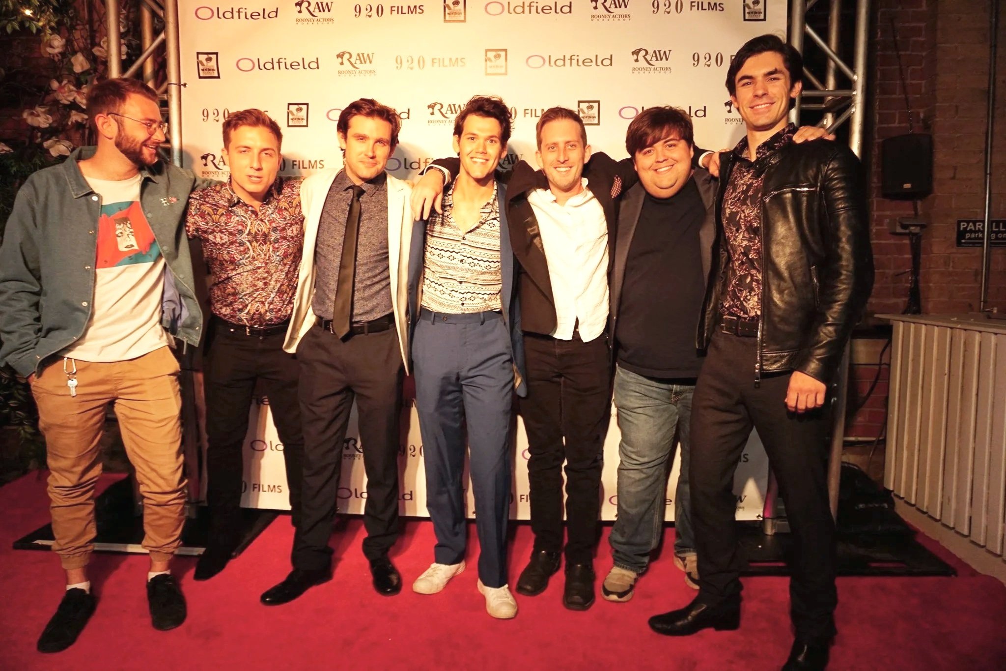 A group of eight young men standing closely together, smiling, at an event with a backdrop displaying 'Oldfield,' 'RAW,' and '920 FILMS' logos. They are in casual and semi-formal attire, with arms around each other's shoulders.