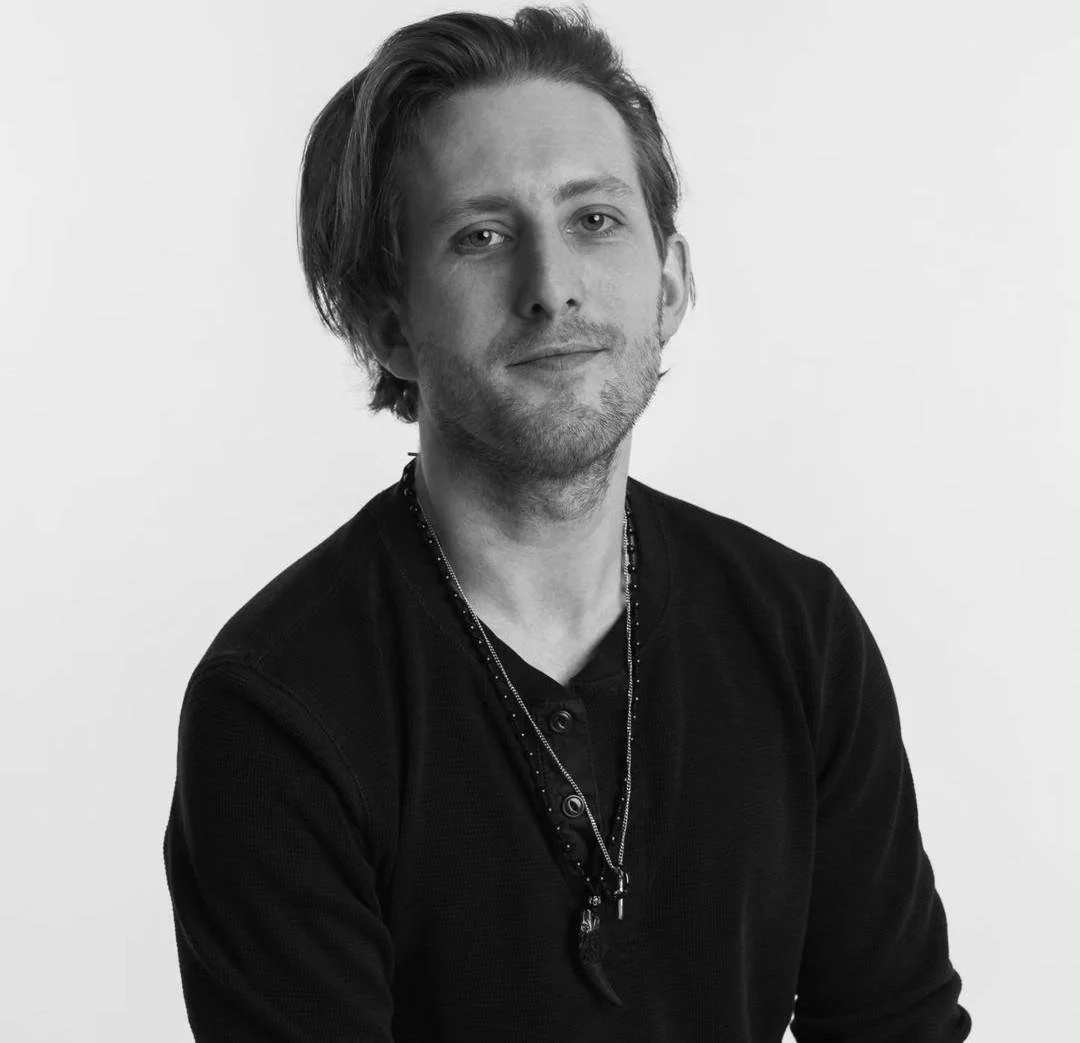 Black-and-white portrait of Michael Joseph Delaney, a young man with medium-length hair, wearing a dark shirt and layered necklaces, looking at the camera with a slight smile.